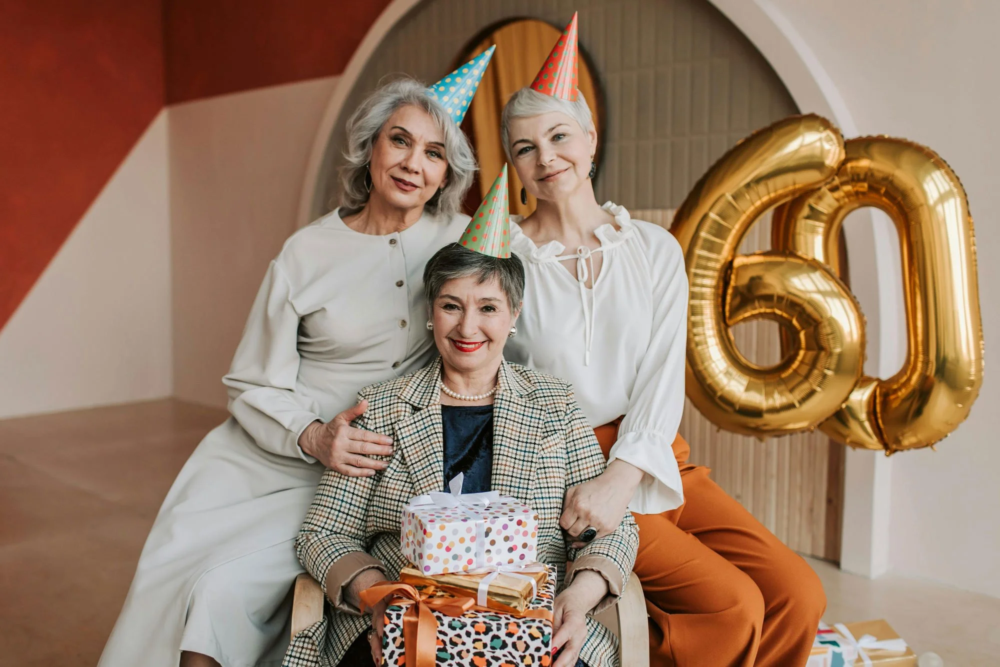 Three women celebrating at a birthday party, wearing party hats, smiling, surrounded by balloons and gifts, with a large golden balloon number 60 in the background.