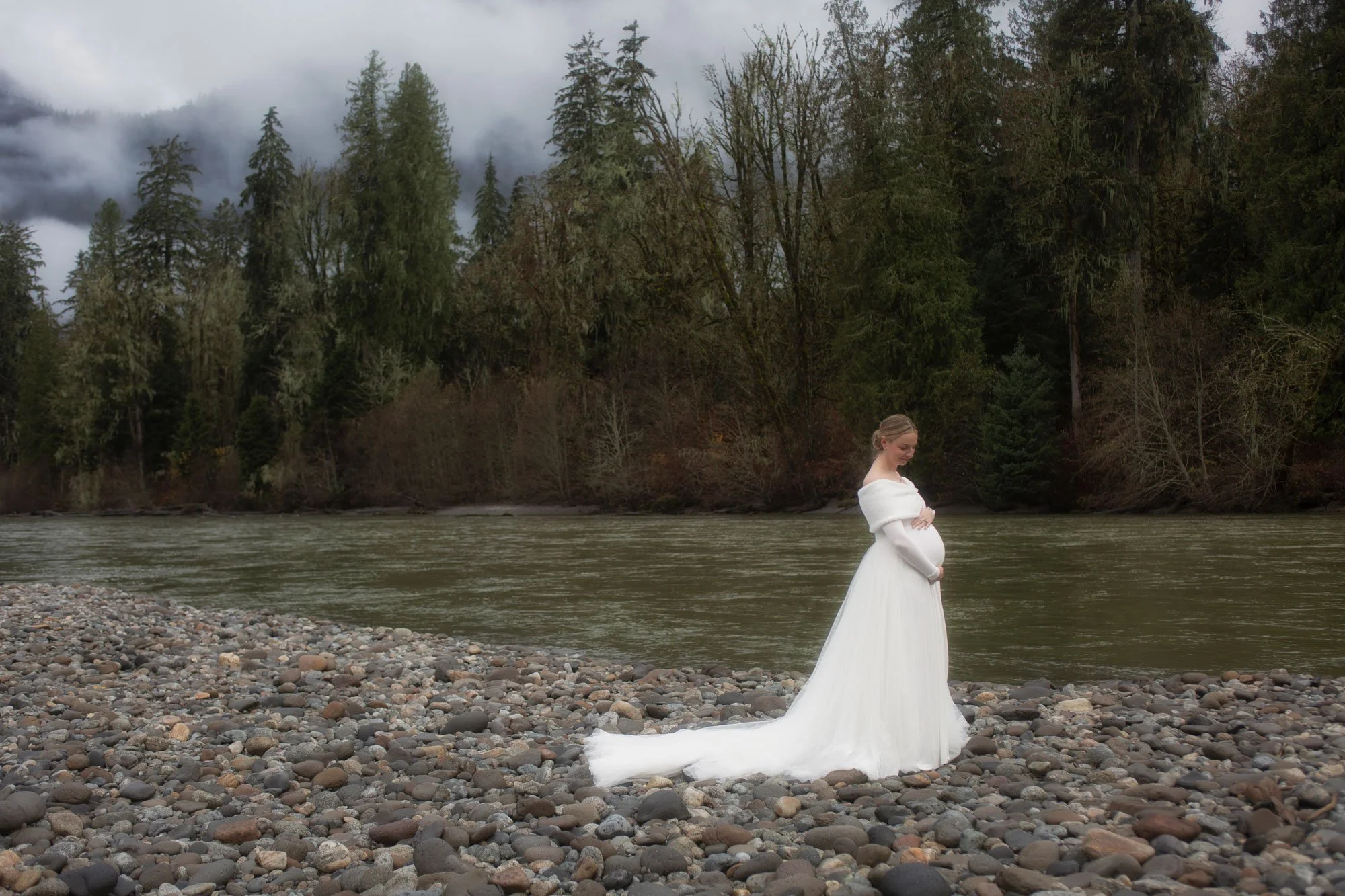 A pregnant woman in a white wedding dress standing on a rocky riverbank with her arms crossed over her belly, surrounded by a forest and overcast sky.