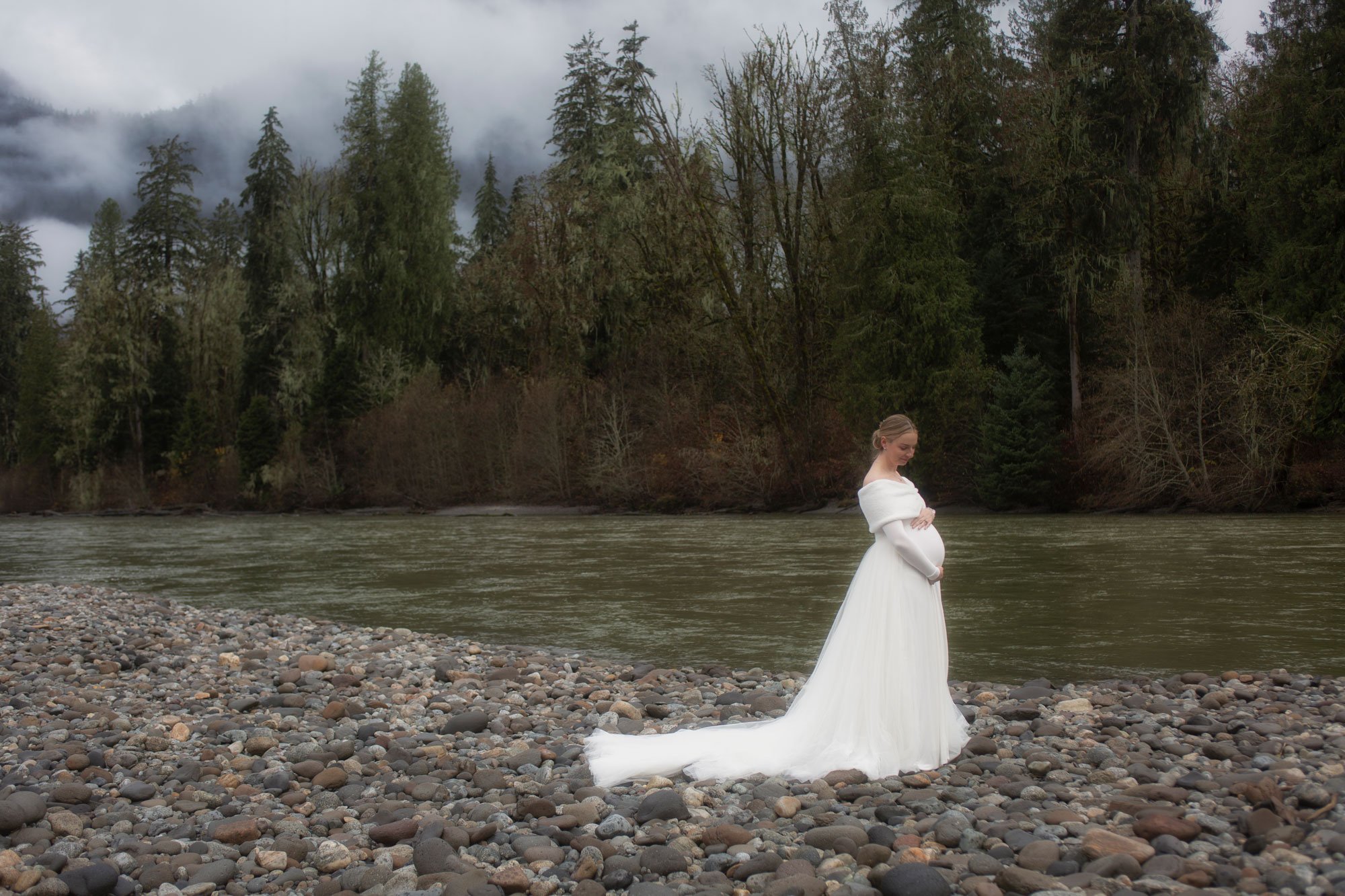 A pregnant woman in a white wedding gown standing on a rocky riverbank with a forest and cloudy sky in the background.