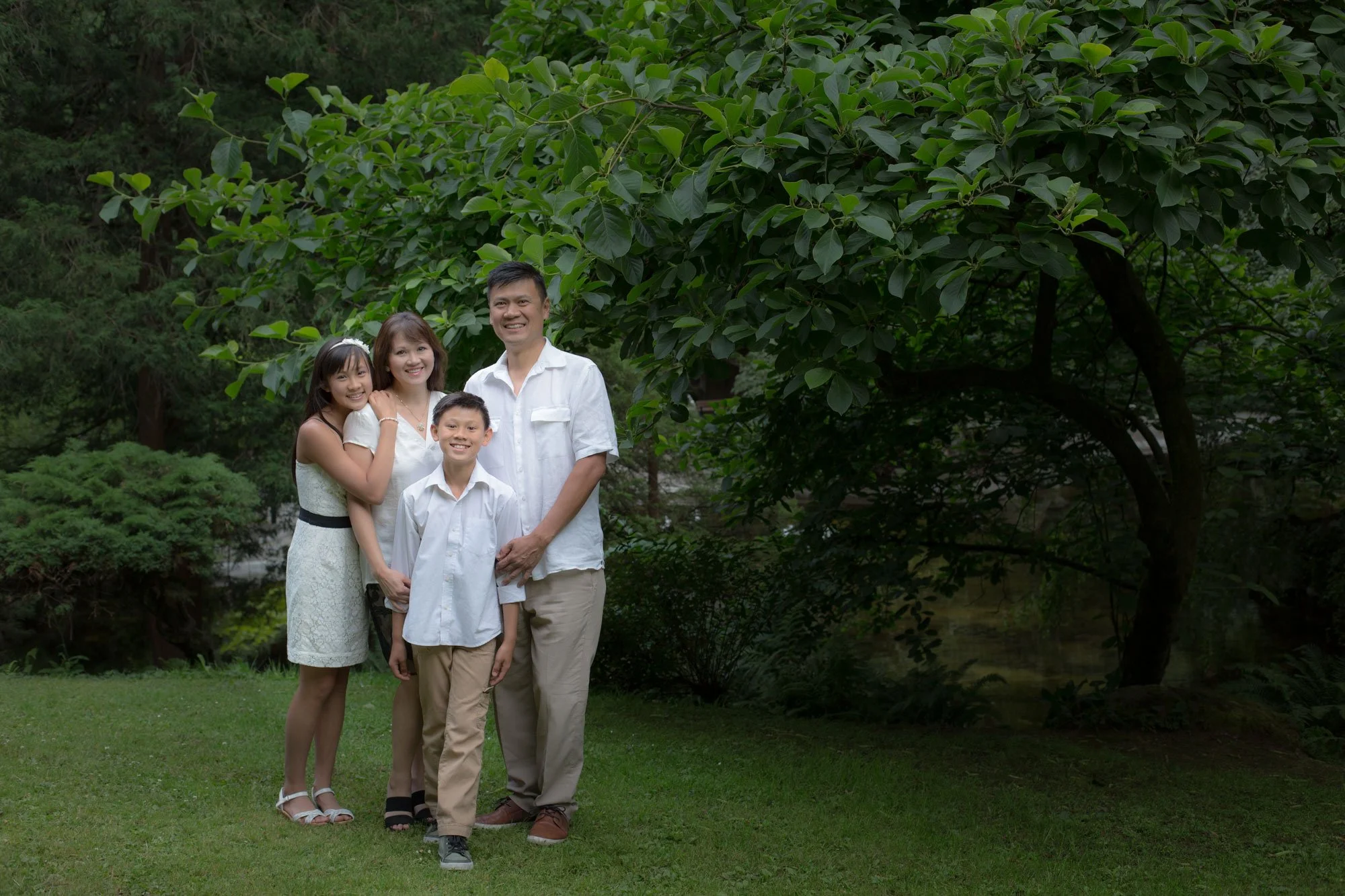 A family of four standing outdoors on grass, smiling, with lush green trees and foliage in the background.