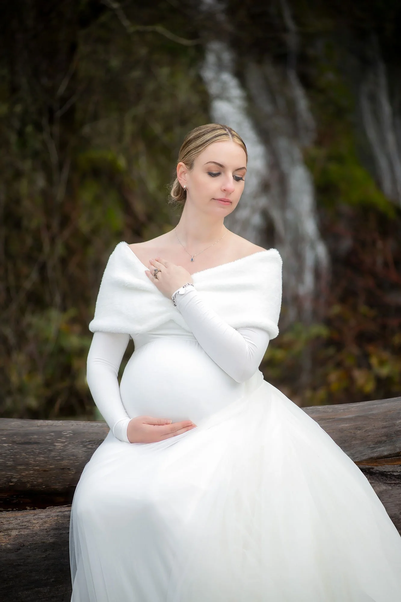 Fine art maternity photograph of a pregnant woman seated on a log with a waterfall in the background, photographed by Andrew Legere.