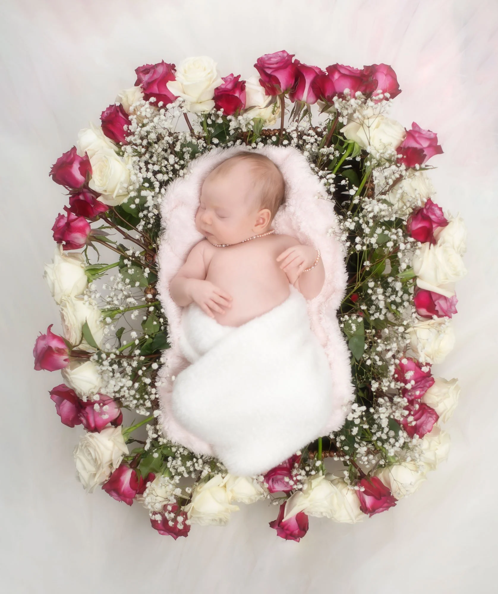 Sleeping baby surrounded by pink and white roses and baby's breath, lying on a pink and white blanket.