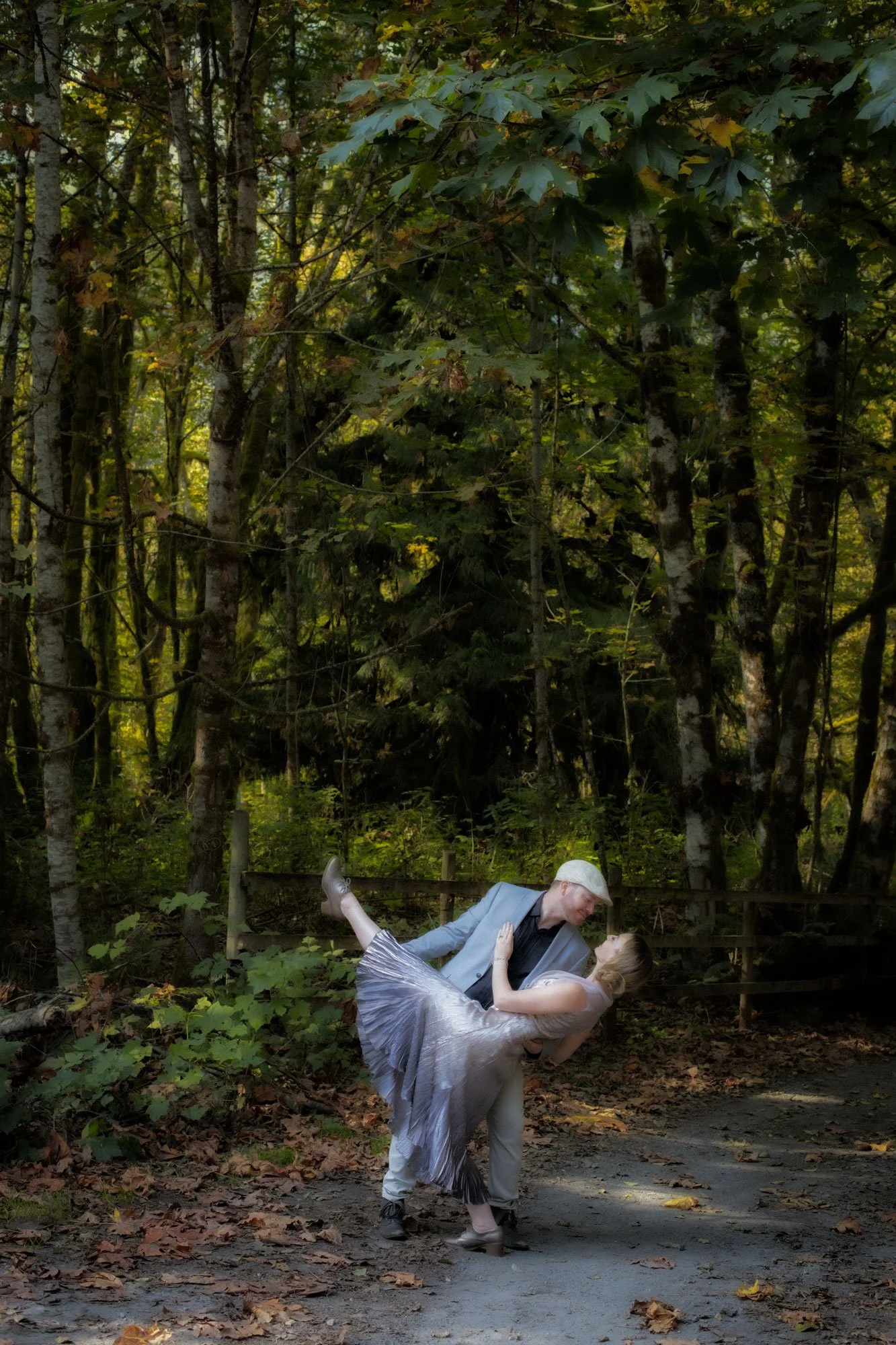 A man in a gray suit and woman in a flowing silver dress dance together outdoors in a wooded area, with the man dipping the woman.