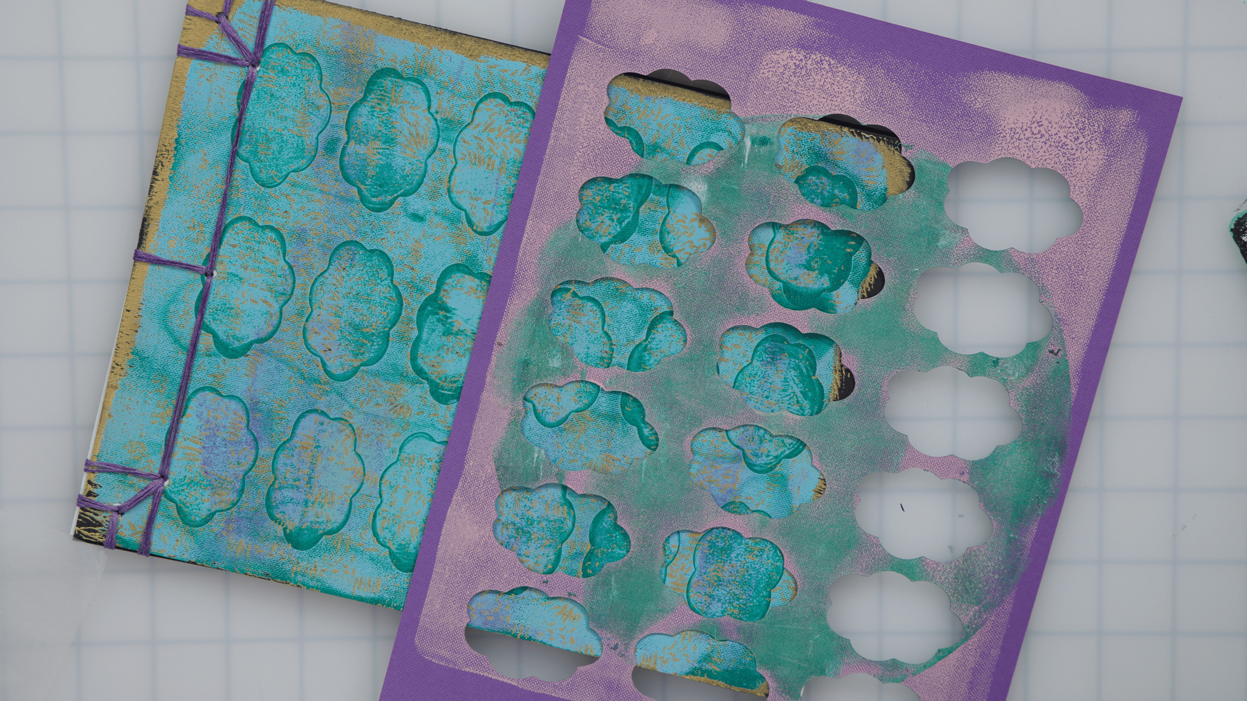 A purple stencil with cloud-shaped cutouts and a teal, purple, and gold patterned sheet underneath. The sheet has cloud-shaped cutouts that match the stencil, and the stencil appears to be used for painting clouds.