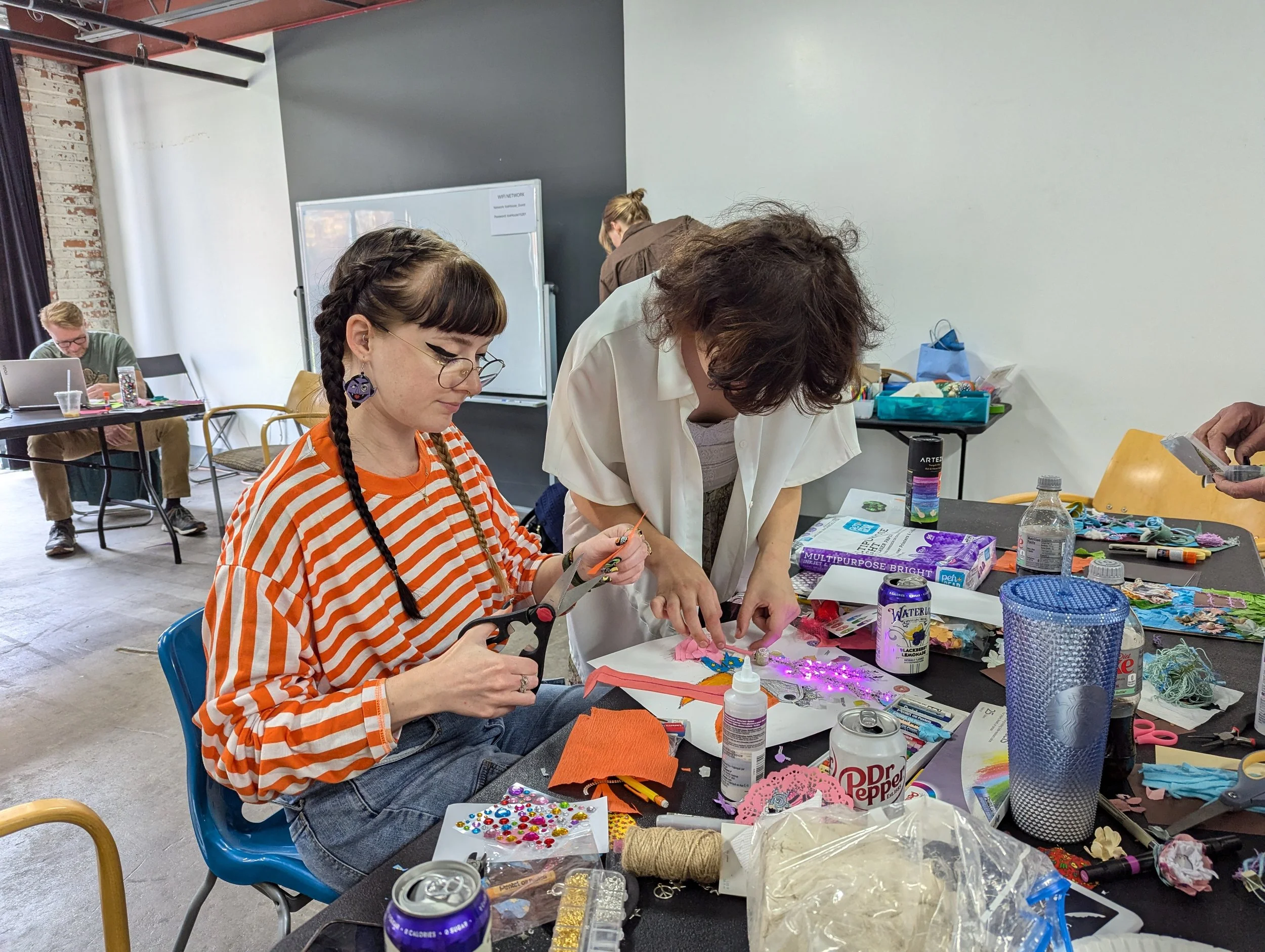 Two women engaged in arts and crafts at a crafting table filled with various supplies, including scissors, glue, beads, and decorative items, in a well-lit room with other people working in the background.
