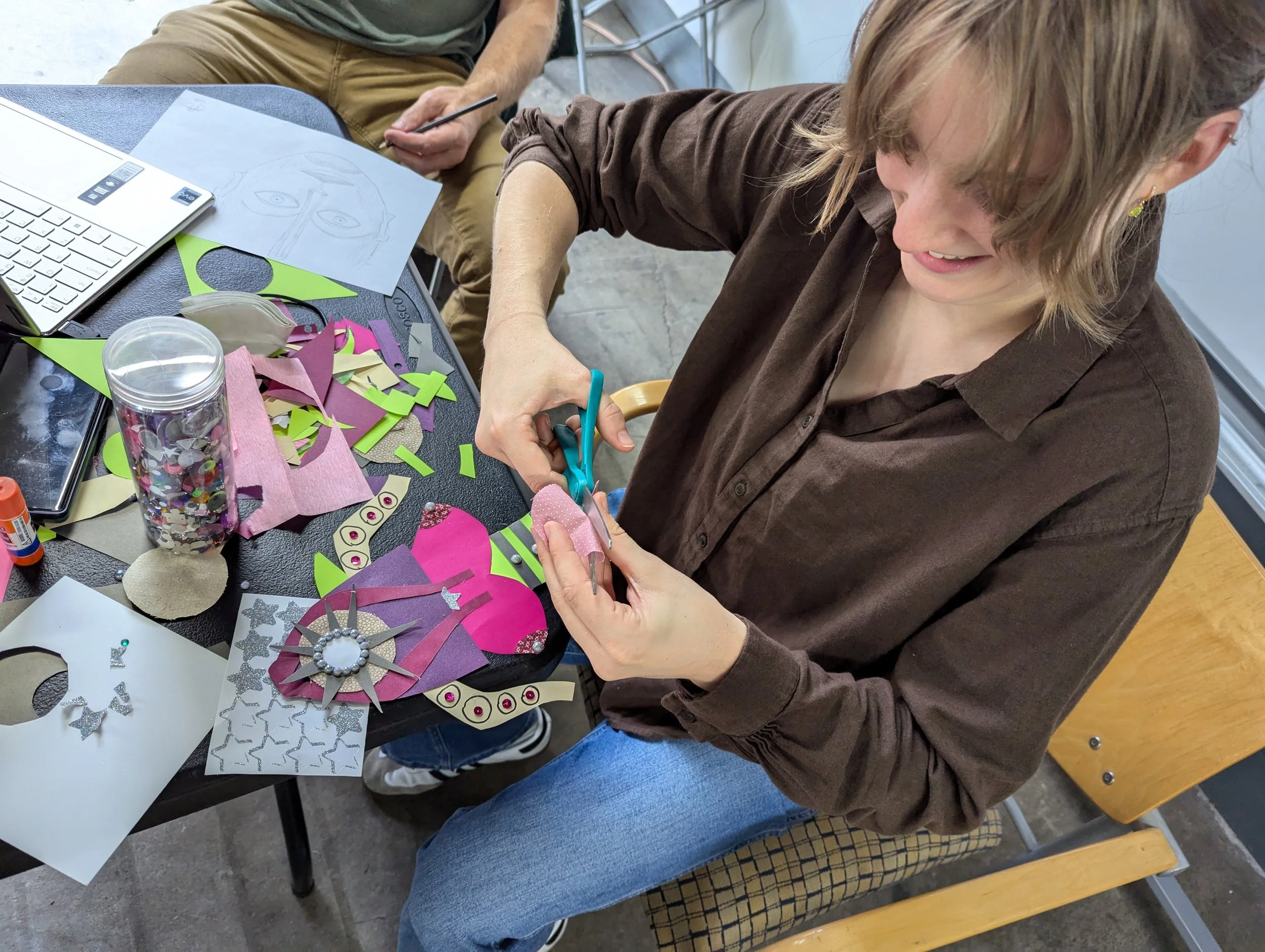 Woman cutting pink and white patterned paper with scissors at a craft table filled with colorful paper, glitter, and craft supplies, while another person sketches on paper in the background.