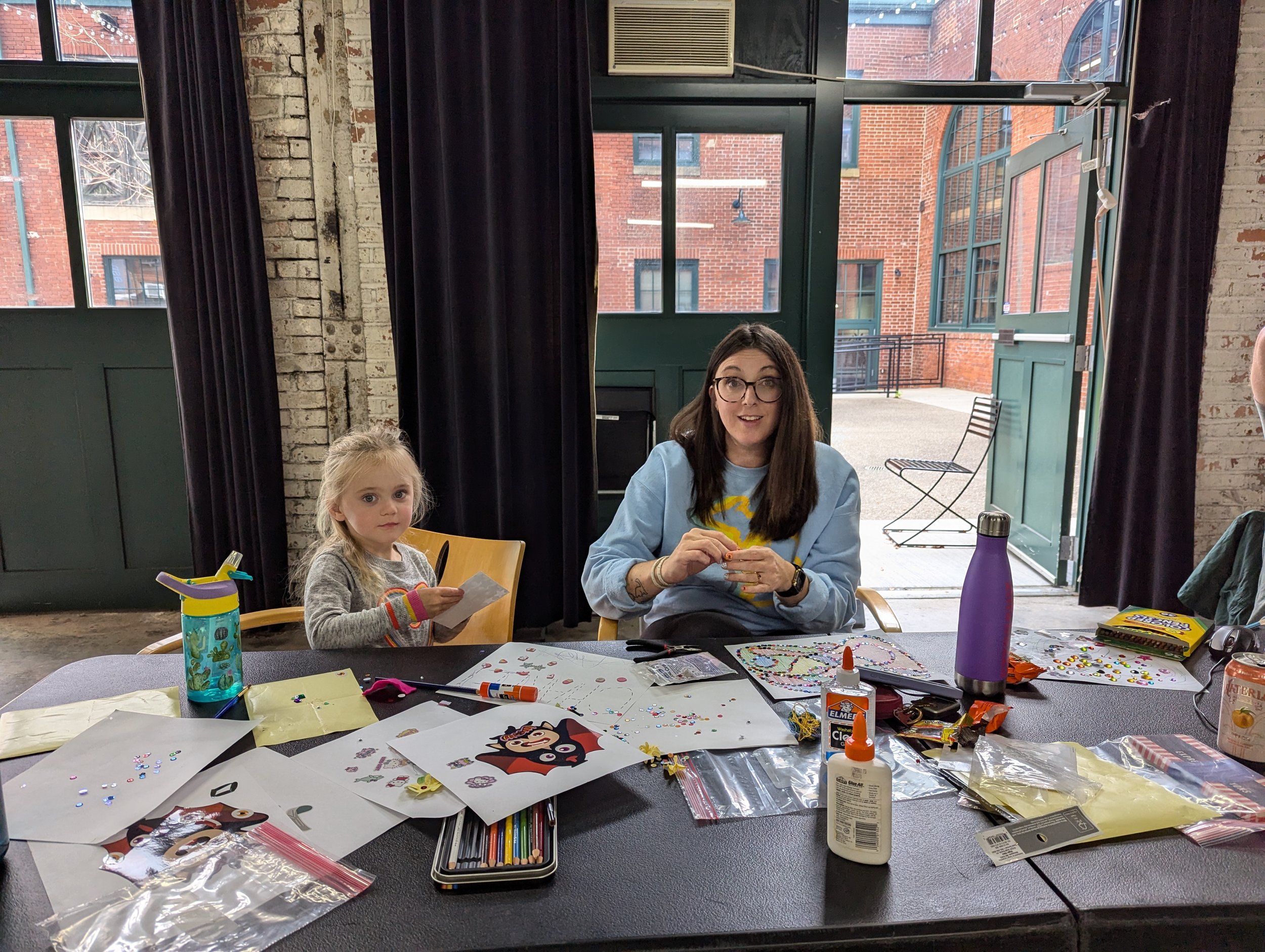 Two females sitting at a table in an indoor room with large windows. The young girl on the left holds a small paper and looks at the camera, while the woman on the right, wearing glasses and a blue sweatshirt, is working on a craft project. The table is filled with craft supplies such as glue, stickers, papers, and drawing tools.