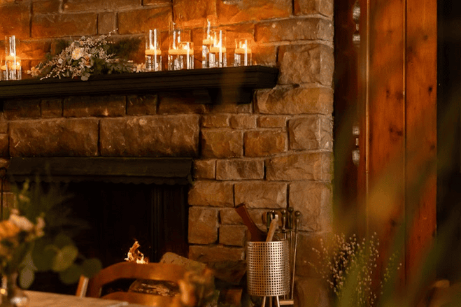 A brick fireplace decorated for the holidays with candles on a mantel, a mix of green foliage and white ornaments, and a black mantel shelf in a cozy room.