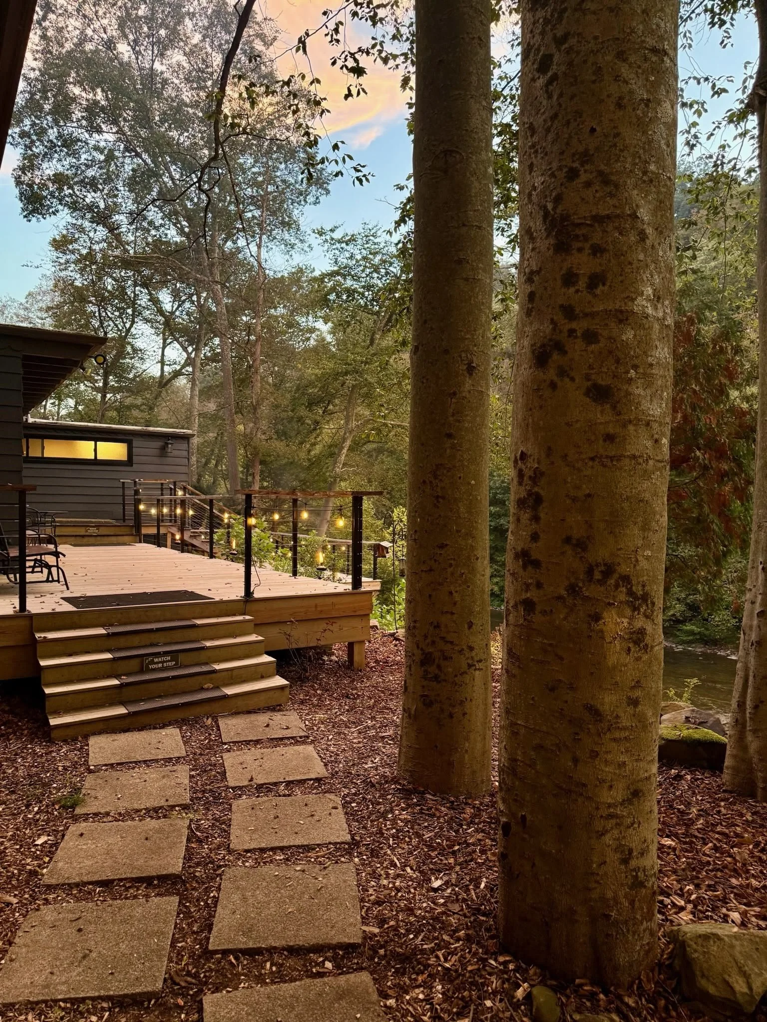 Wooden deck with string lights, steps leading up to deck, surrounded by trees and forest, with a river nearby, during sunset.