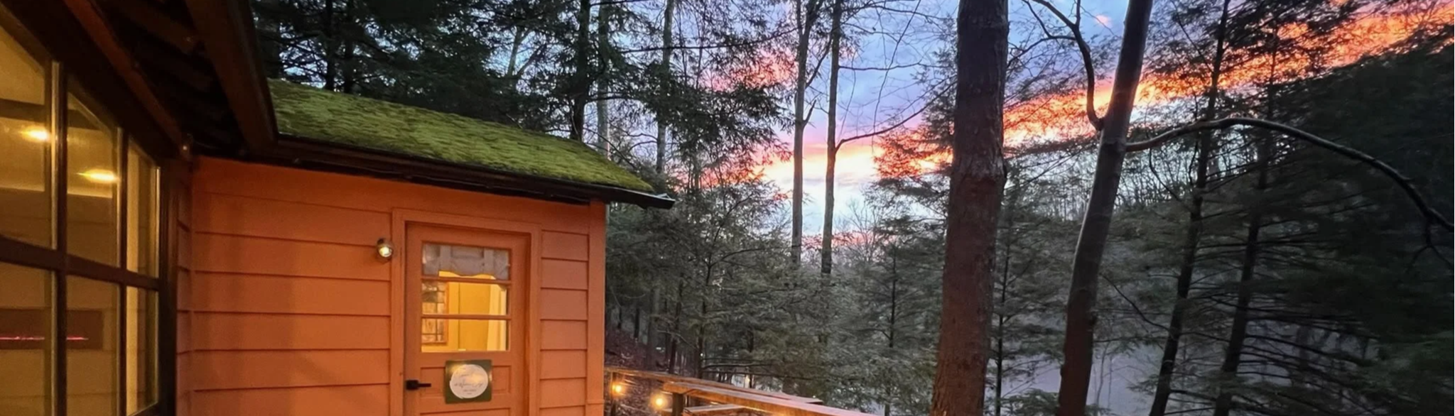 Sunset sky over a forest viewed from a cabin, with a corner of the cabin's orange wall and window illuminated inside.