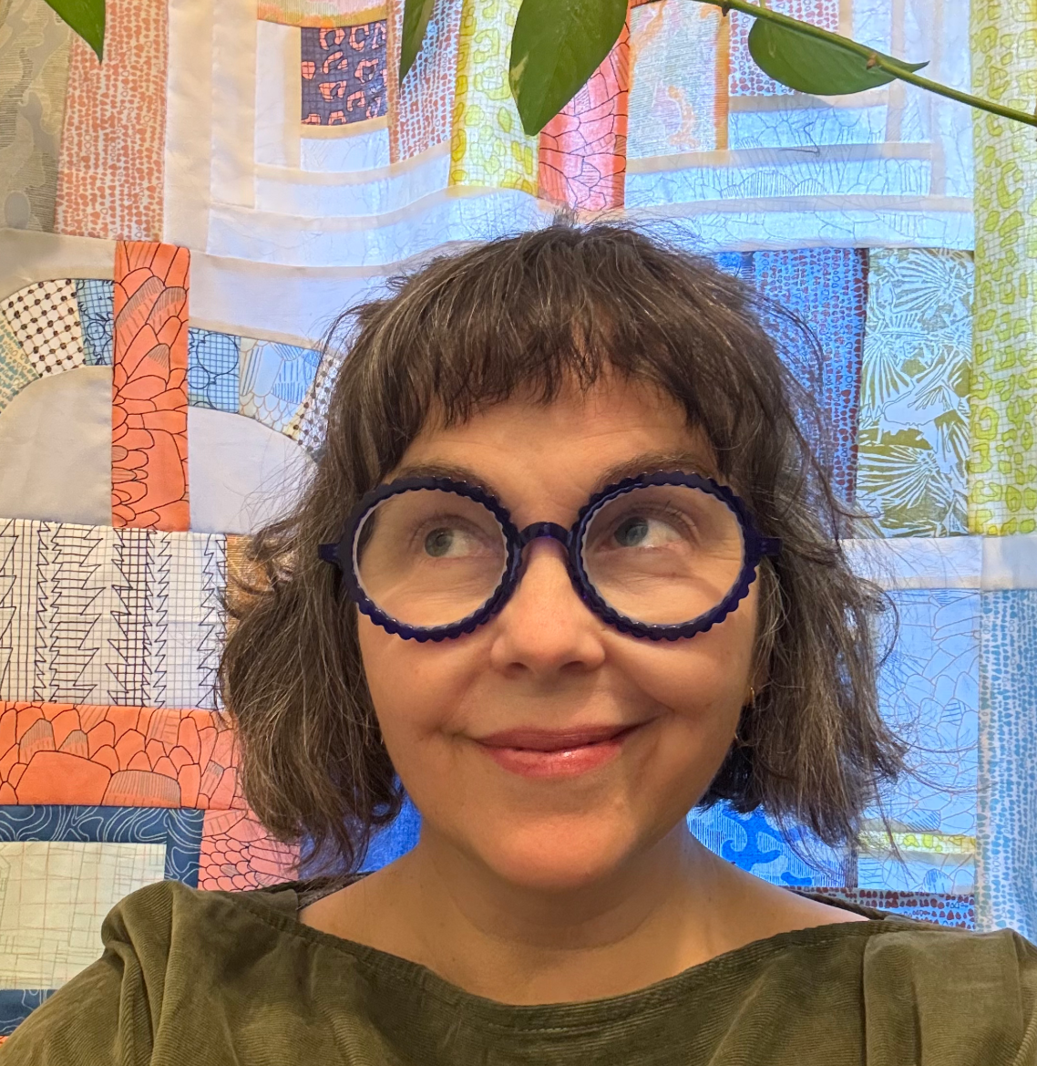 A woman with short brown hair and large round dark purple glasses looking slightly upwards with a playful smile. She is standing in front of a colorful patchwork quilt with various patterns and colors, including orange, blue, pink, and green.