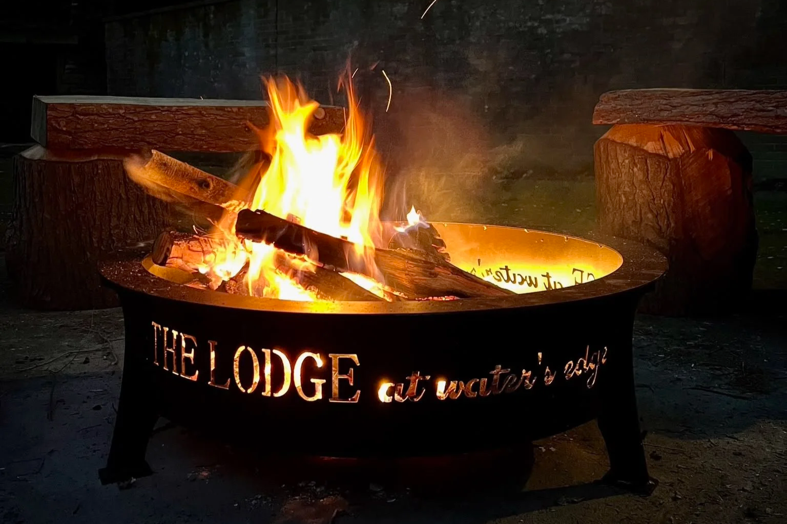 A lit outdoor fire pit with the words 'THE LODGE at water's edge' cut out from the side, surrounded by logs, with flames burning and wood inside.