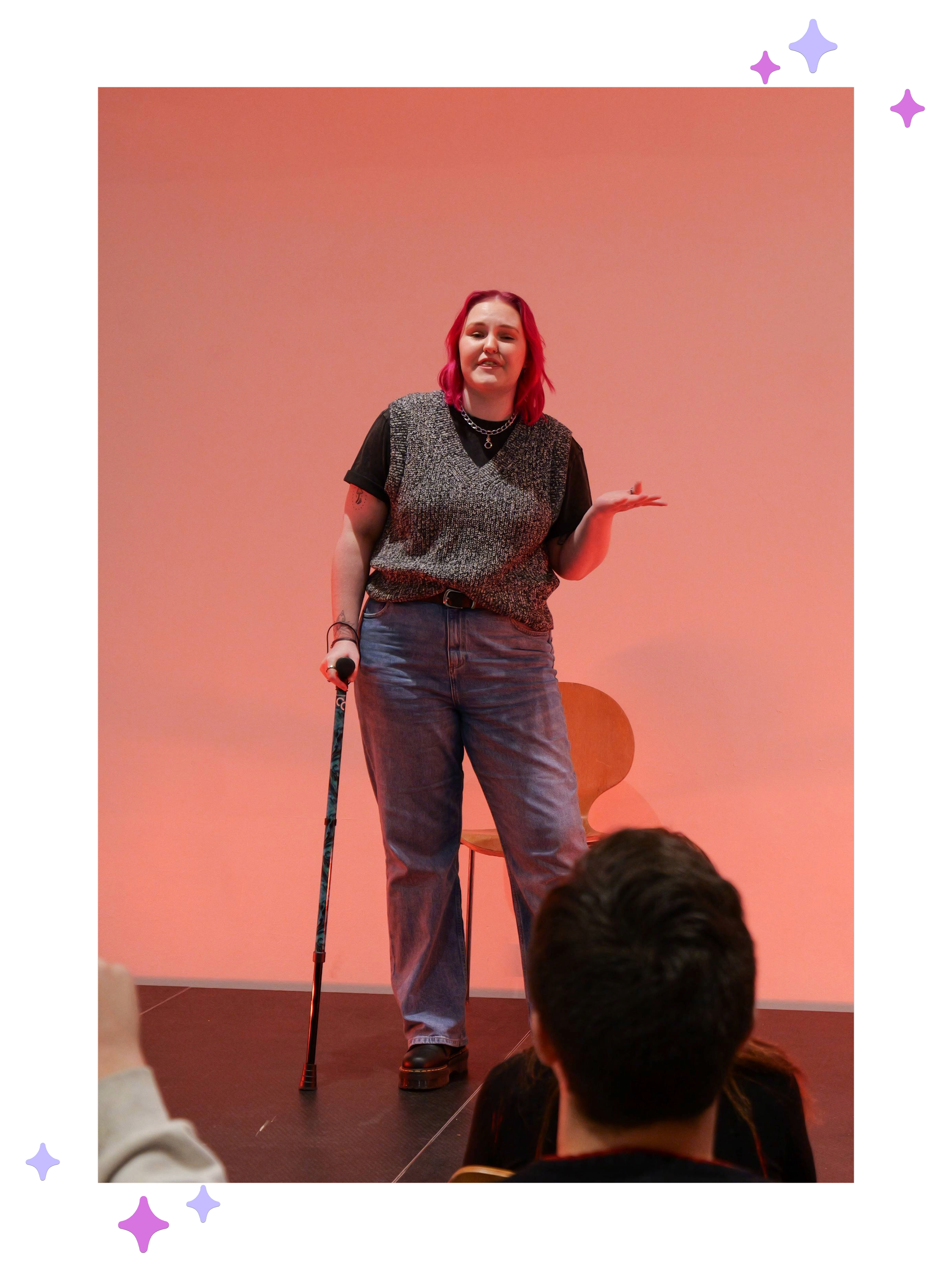 Lozza, a white non-binary person with pink hair stood on a small stage delivering a monologue. The back wall is lit up orange and they’re disabled and using a black and green stick