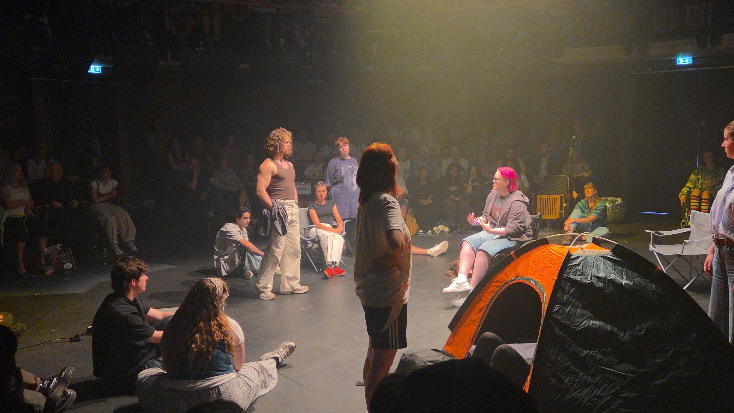 A group of theatre performers on stage in the round at Rose Bruford college. Lozza is sat in their chair performing almost centre stage while the other actors watch