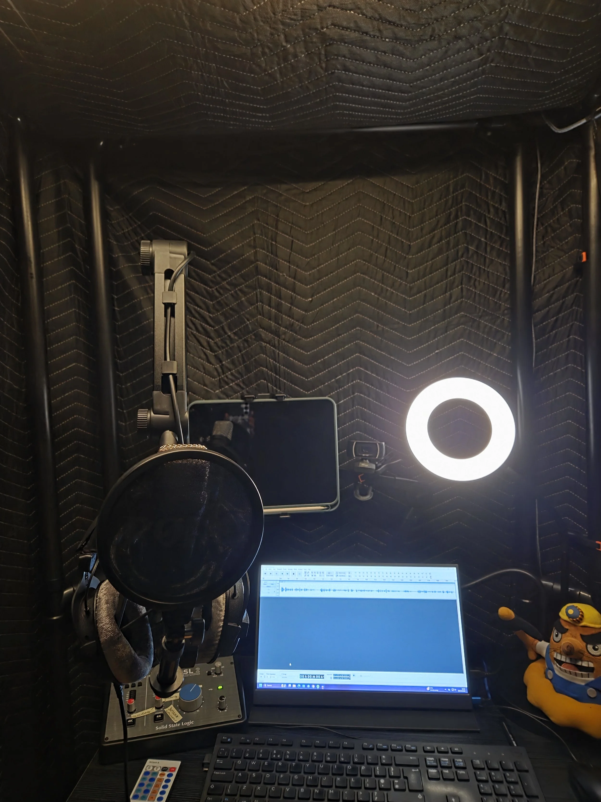 Lozza’s home studio. It is a pvc frame with sound blankets hung over it. Inside there is a Rode NT1 microphone on a boom arm with a pop filter attached and Beyerdynamic DT 770 Pro headphones hung on it, a small desk, a monitor with webcam, ring light
