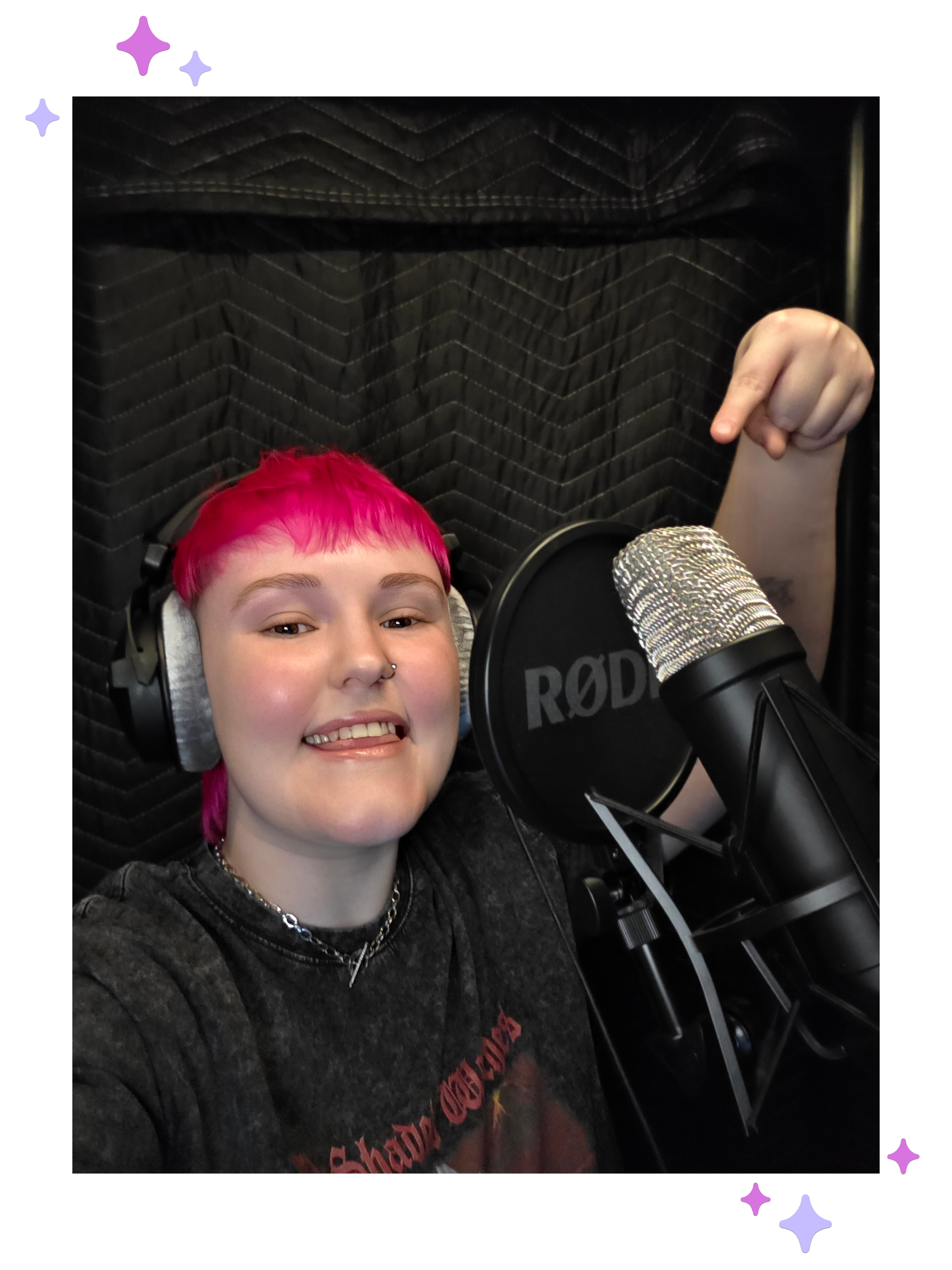 Lozza, a white non-binary person with a bright pink mullet in their voiceover booth. They’re pointing at their Rode microphone and smiling while wearing headphones