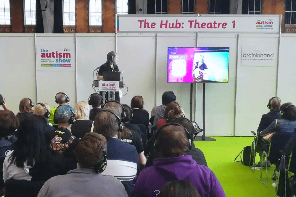 Lozza turning to their final slide of their talk at the Autism Show to say thank you and answer questions. They're stood/sat on a small stage in front of a podium with a laptop on it and the full audience is listening via headphones.