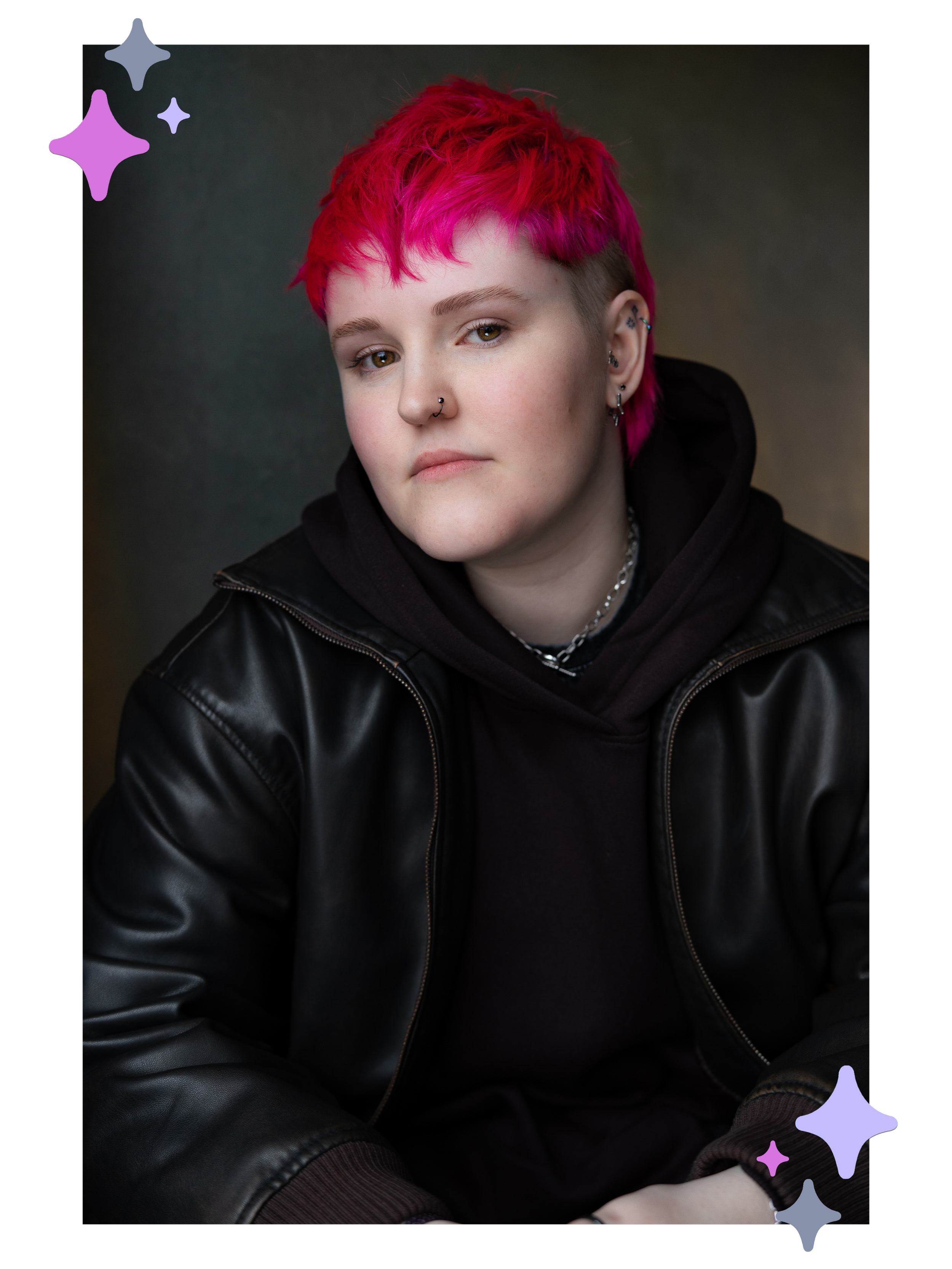 Headshot of Lozza, a white non-binary person with a bright pink mullet wearing a leather jacket with a black hoodie under it. They’re looking at the camera almost side-on with a neutral expression