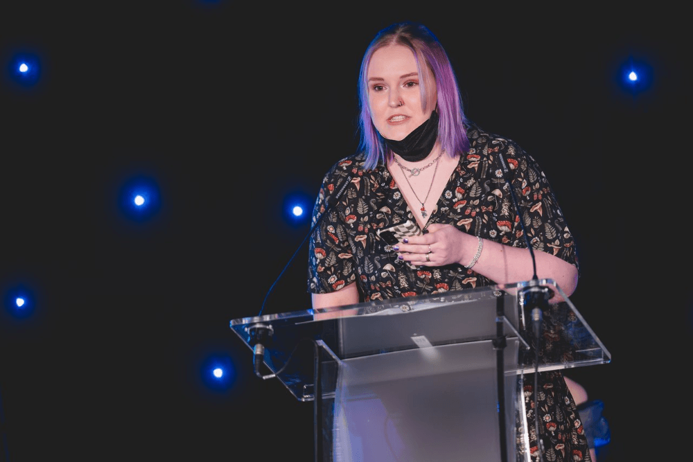 Lozza, a white non-binary person with purple hair passionately delivering an awards  acceptance speech on stage. They have lowered their mask and are wearing a dress  with mushrooms on it.