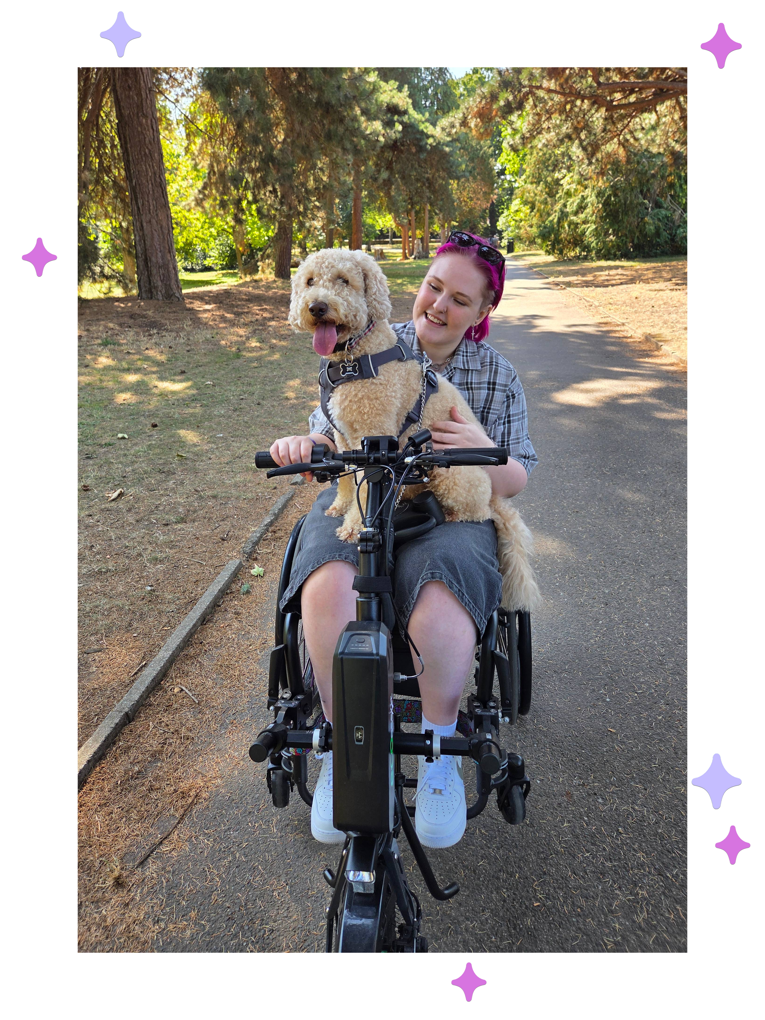 Lozza, a white non-binary person with pink hair sat in their wheelchair in a park with their dog Barney sat on their knee, they’re looking at him and smiling