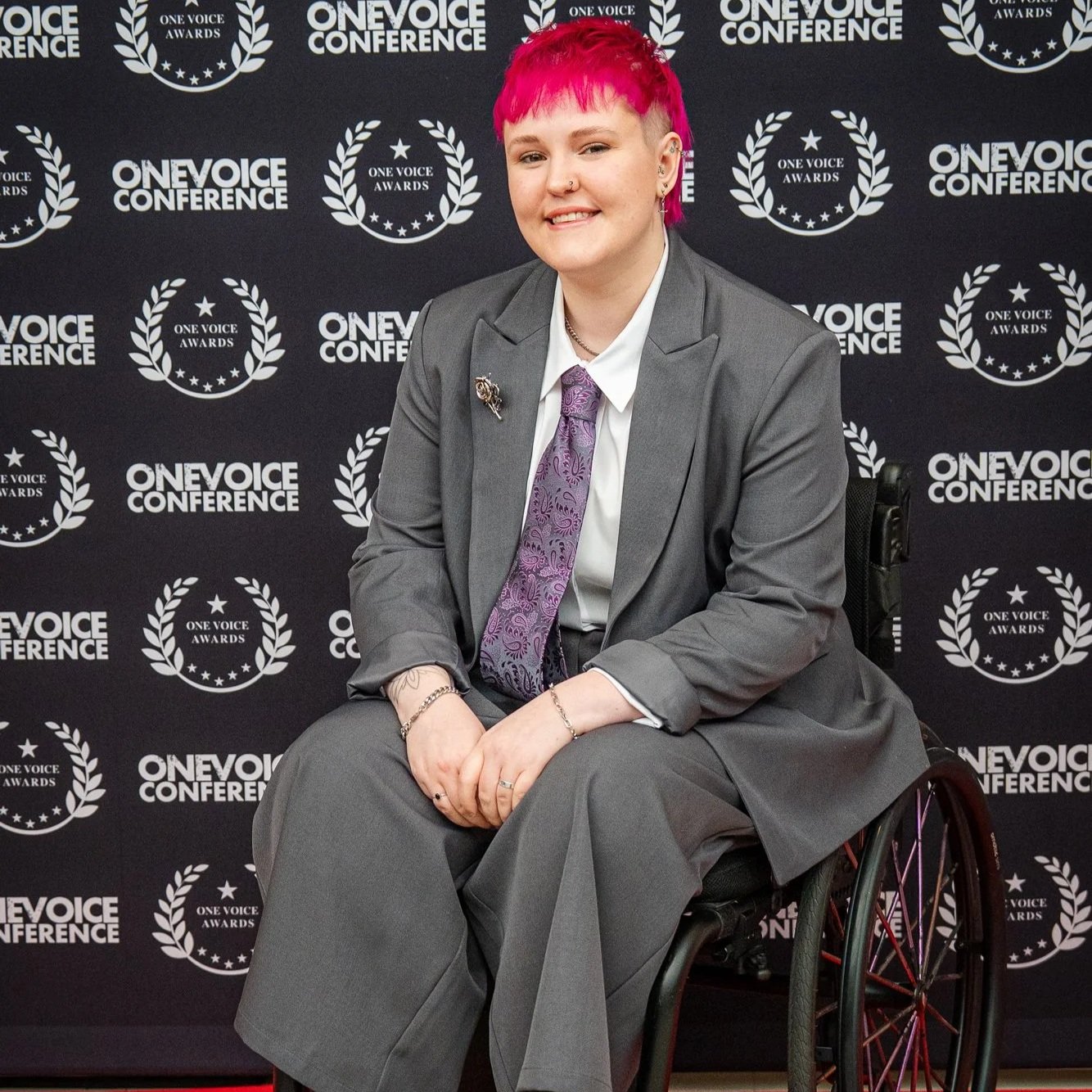 Lozza, a white non-binary person and wheelchair user on the 'red carpet' for the One Voice Awards. They have a bright pink mullet and are wearing a grey suit with a matching tie that has purple details on it. They are smiling at the camera.