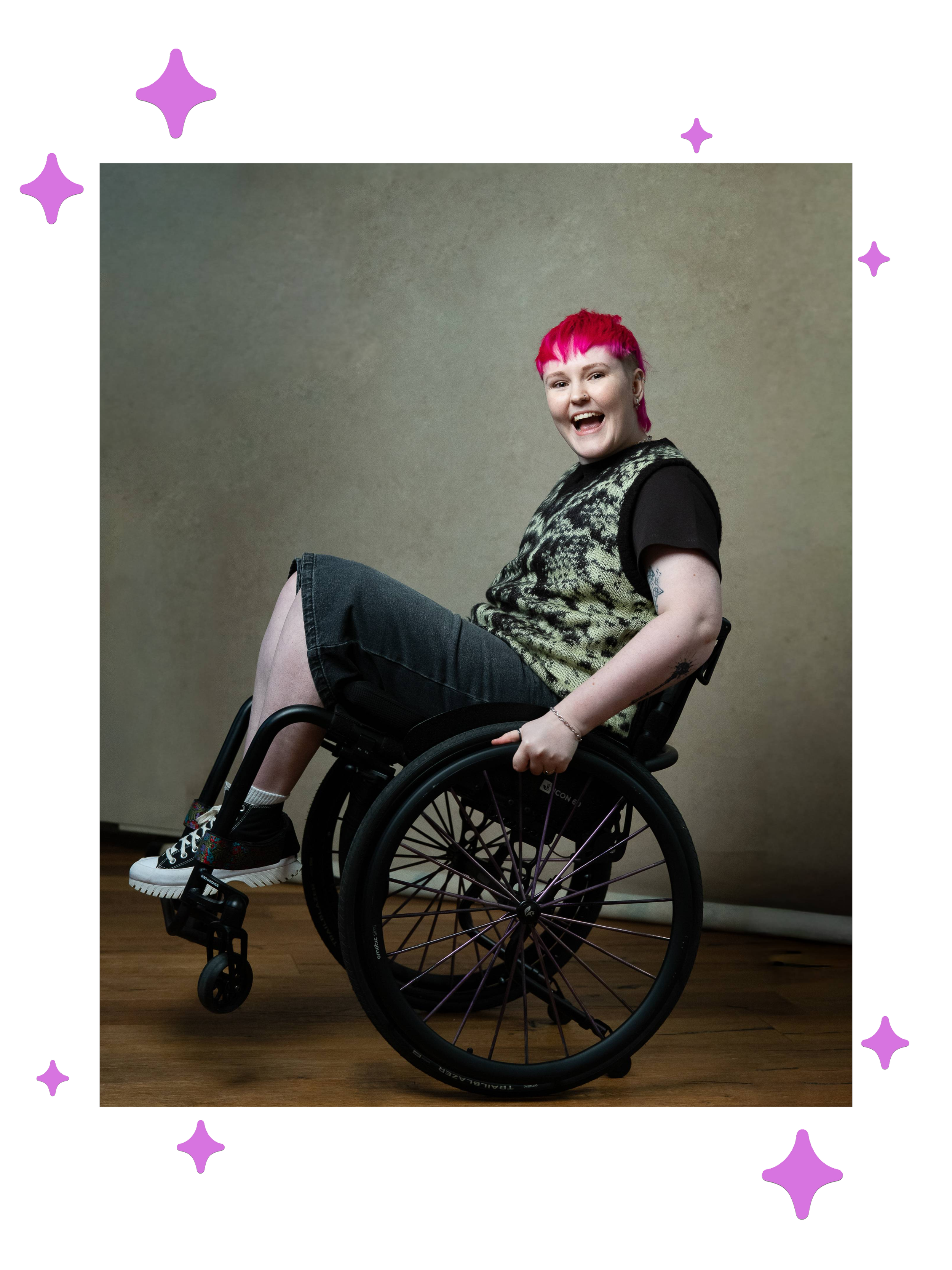 Lozza, a white non-binary person with a bright pink mullet pulling a wheelie in their wheelchair. They’re wearing black jorts with a black t shirt and a black and green sweater vest and looking at the camera excitedly with their mouth open.