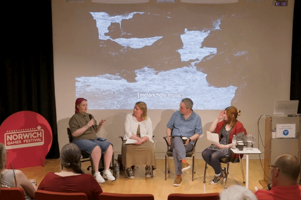 Lozza, Katie, Brendon and Jess speaking on a panel at Norwich Games Festival about Rez High. There's a projector screen behind showing the trailer, a pink Norwich Games Festival sign to the left, and Lozza is speaking with a mic.