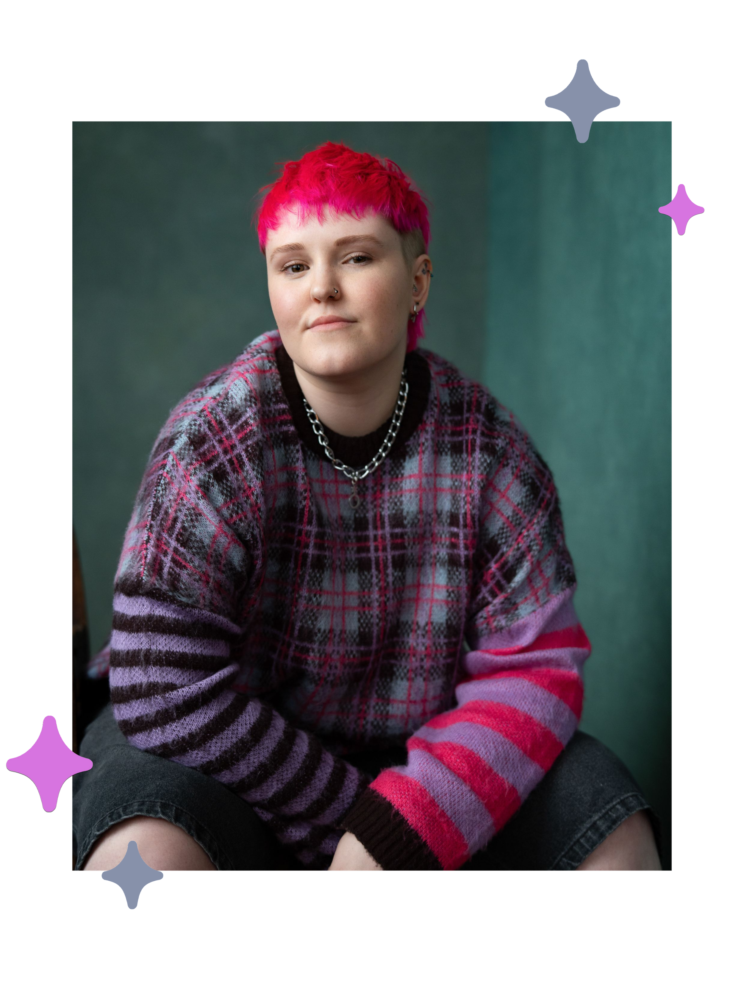 Lozza, a white non-binary person with a bright pink mullet sat with their elbows resting on their knees. They’re wearing a purple, pink and black tartan jumper with black jorts and a silver chain necklace. They’re looking at the camera cheekily