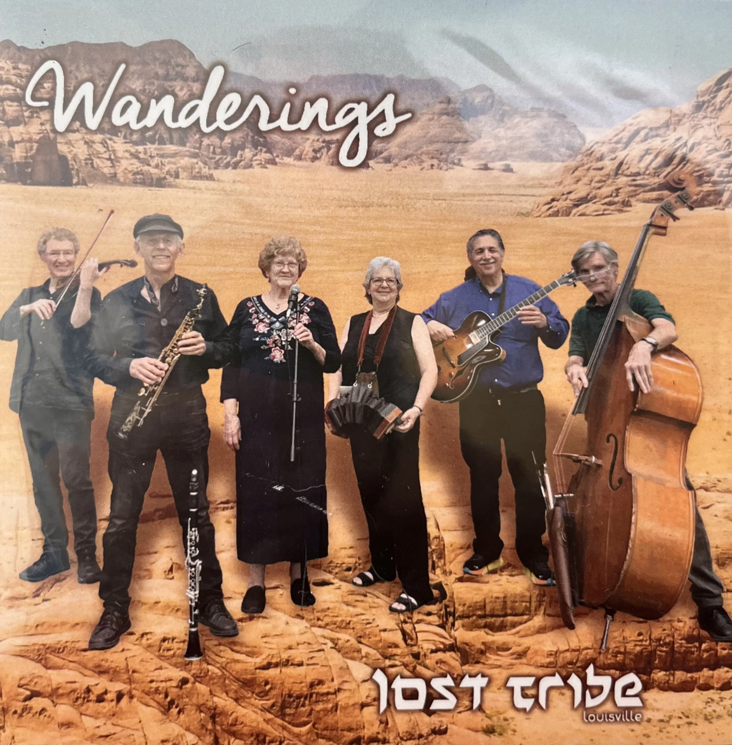 A band of six musicians performing in front of a desert mountain landscape, titled 'Wanderings' by Lost Tribe Louisville.