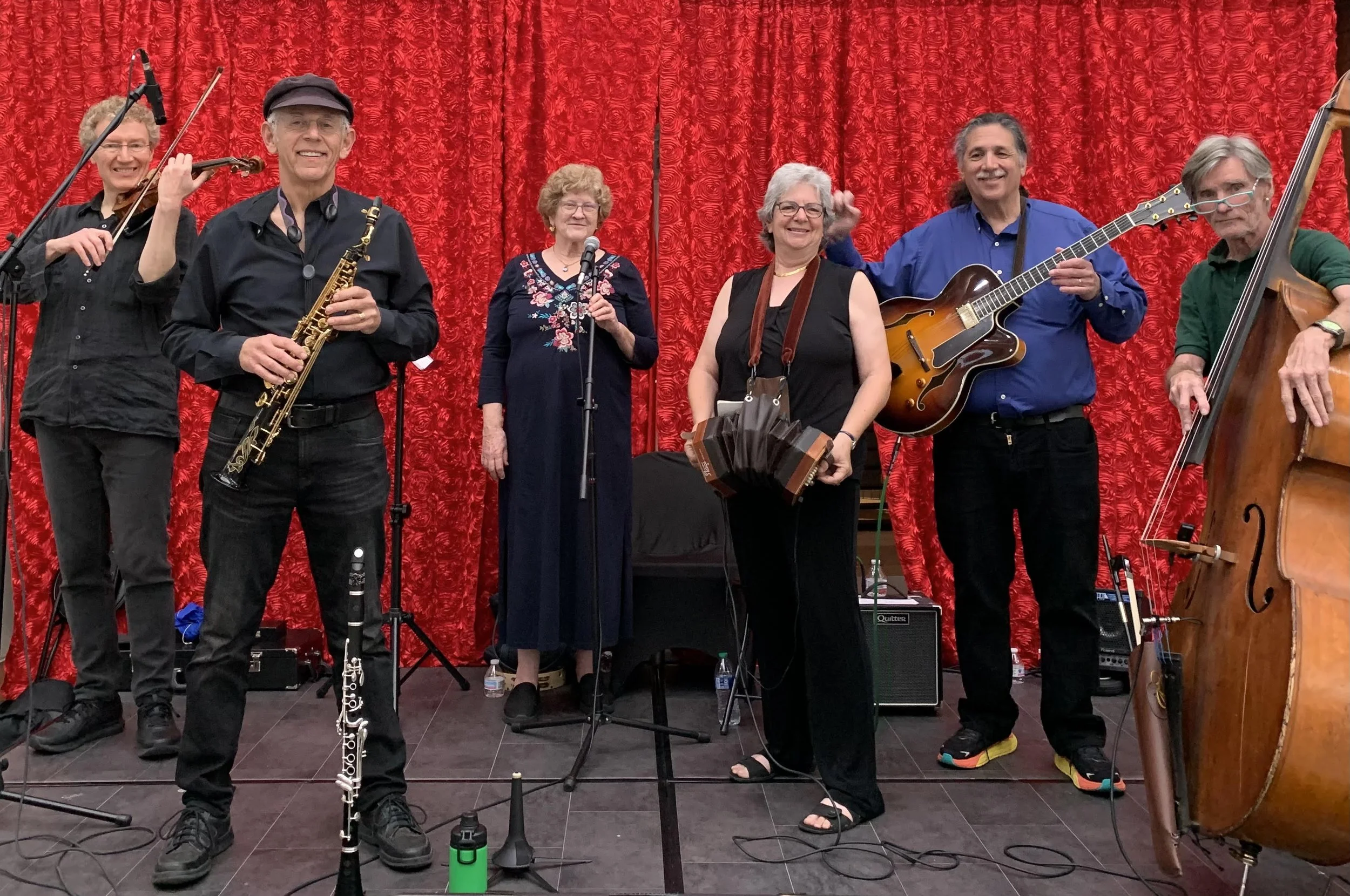 Group of seven musicians performing on stage with red curtain backdrop, holding instruments including a saxophone, guitar, double bass, and microphone.