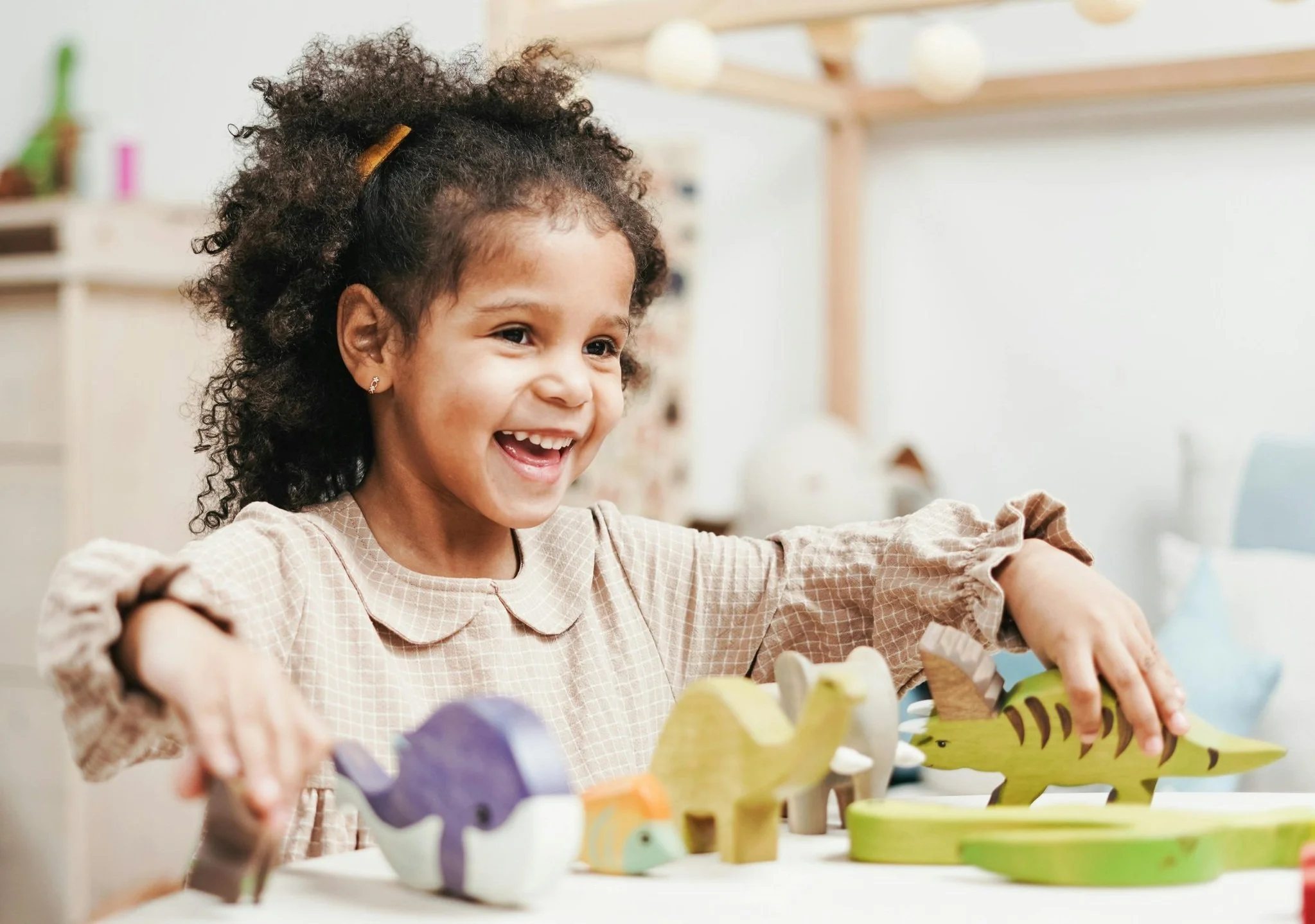 A young girl with curly hair smiling and playing with colorful dinosaur-shaped toys on a white table in a bright room.
