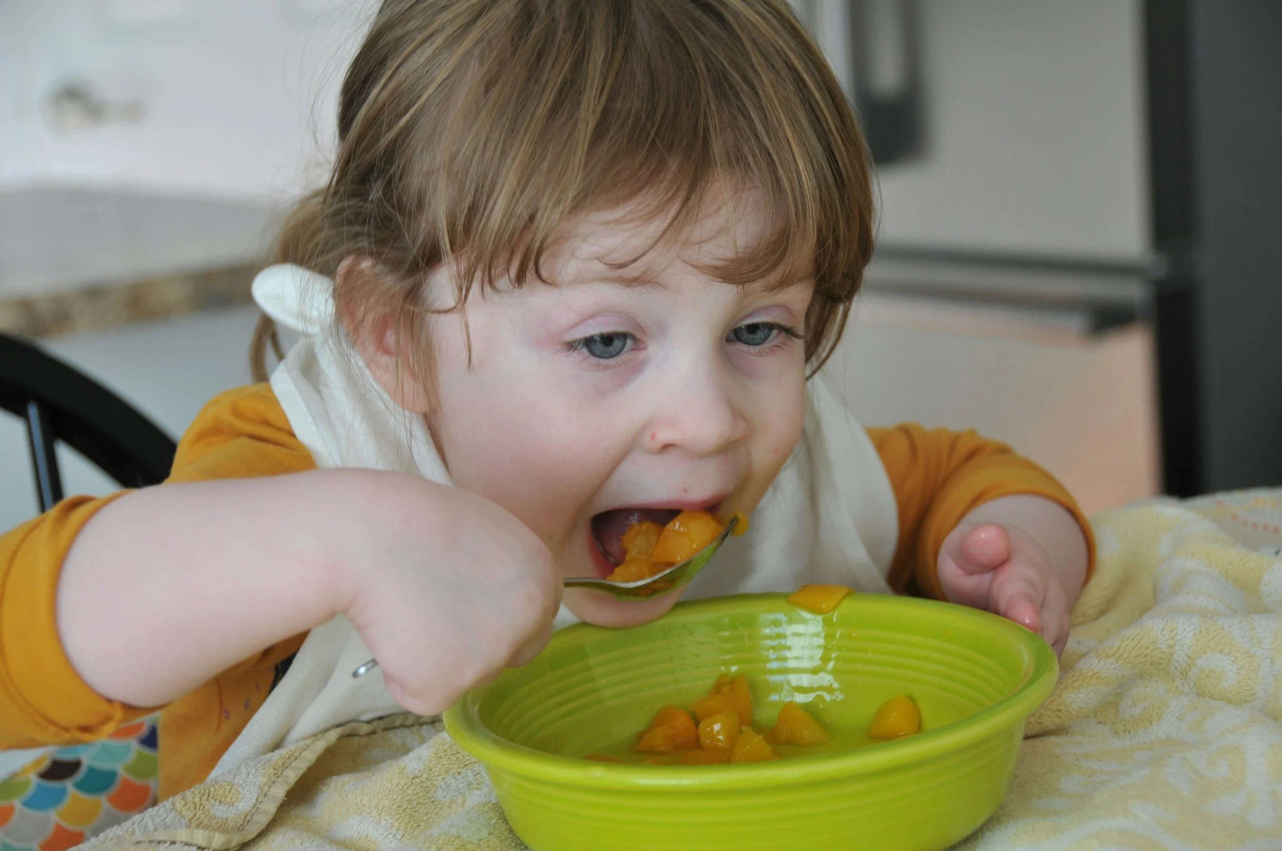 A young child with light brown hair and blue eyes, wearing an orange long sleeve shirt and a white bib, eating from a green bowl filled with chopped peaches using a spoon.