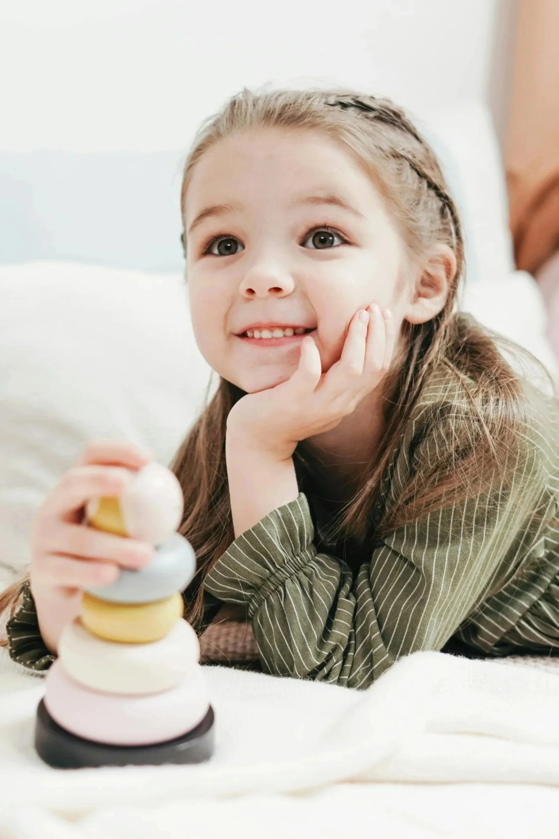 Young girl with brown hair lying on her stomach, resting her face on her hand, smiling, next to a colorful stacking toy.