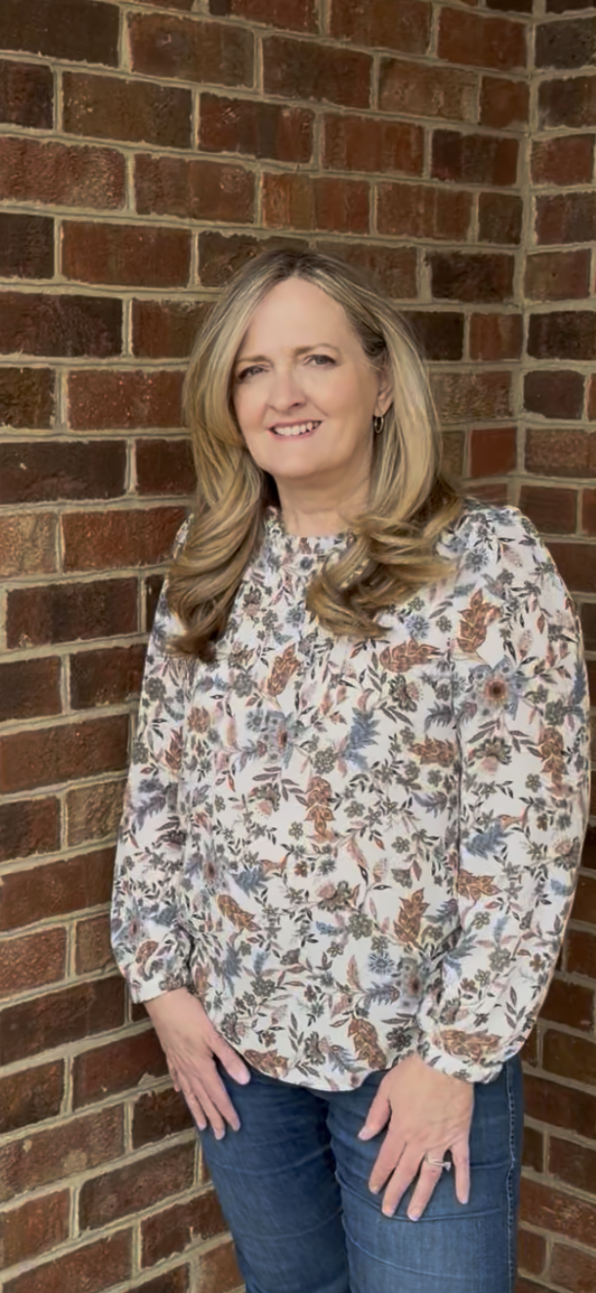 A middle-aged woman with blonde, wavy hair stands near a brick wall, wearing a floral long-sleeve blouse and jeans, smiling at the camera.