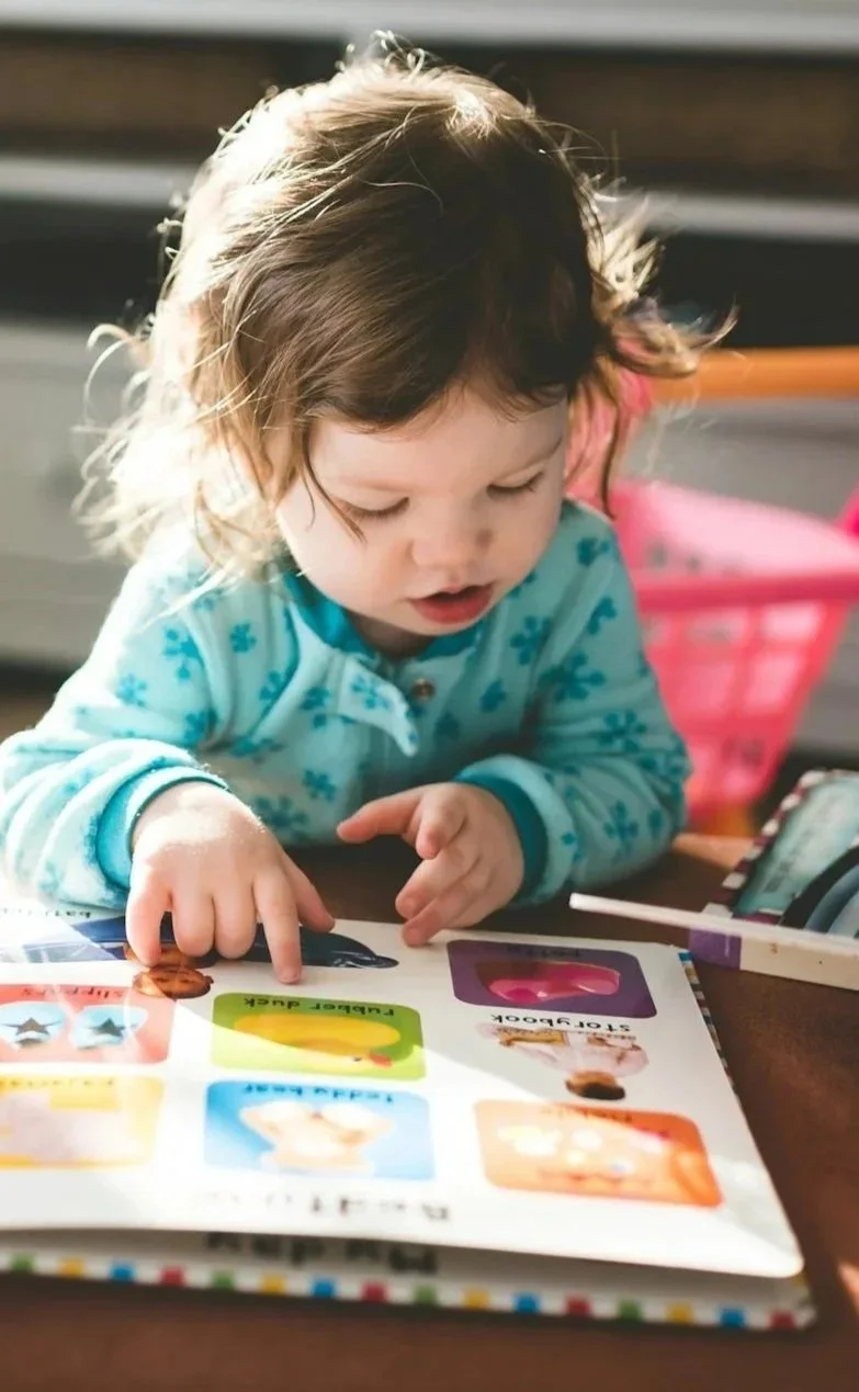A young child with light brown hair looking at a colorful book with pictures of superheroes on a table.