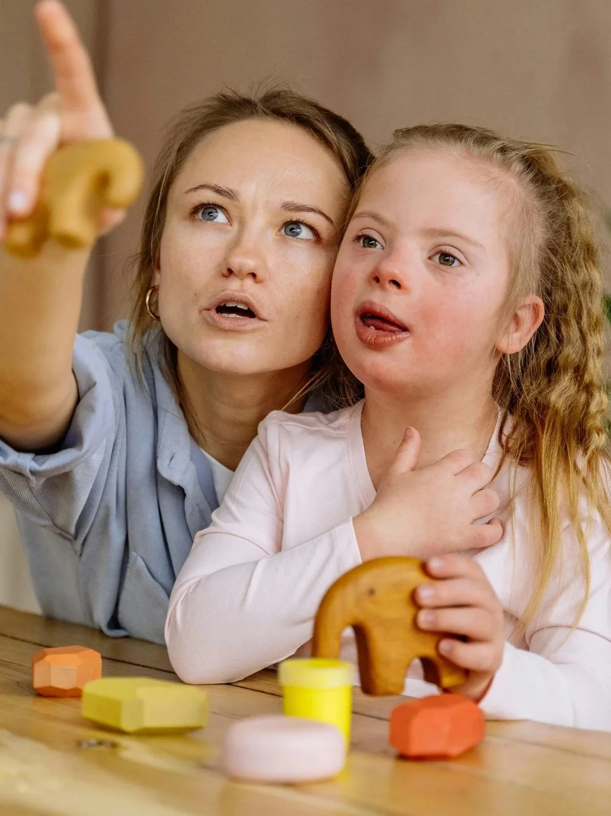 An adult woman and a young girl are playing a game at a table with colorful wooden blocks. The woman is pointing and explaining something, while the girl seems to be attentively listening with her hand on her neck.