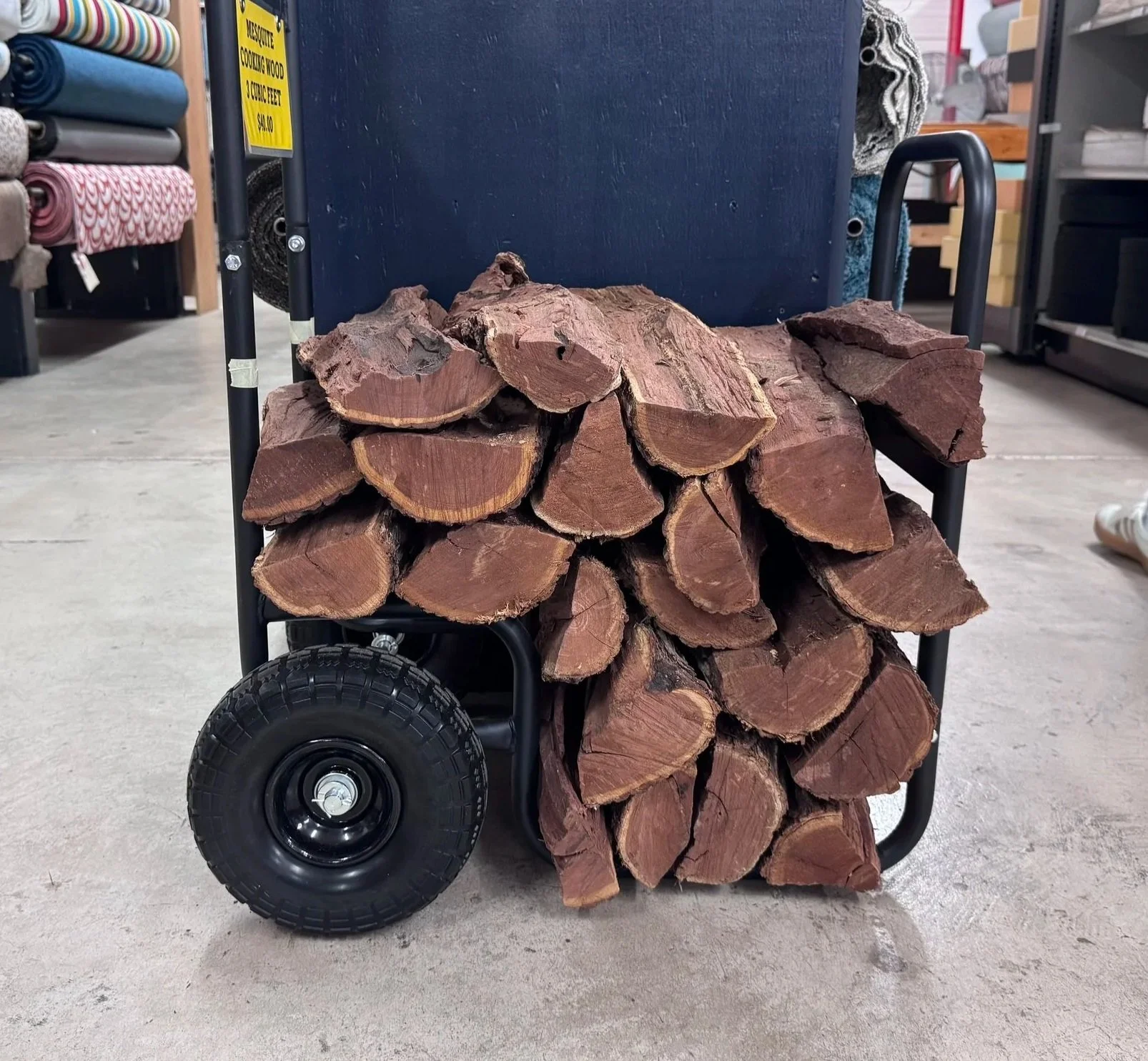 A cart loaded with cut firewood pieces, placed on a store floor with shelves and rolled fabric in the background.