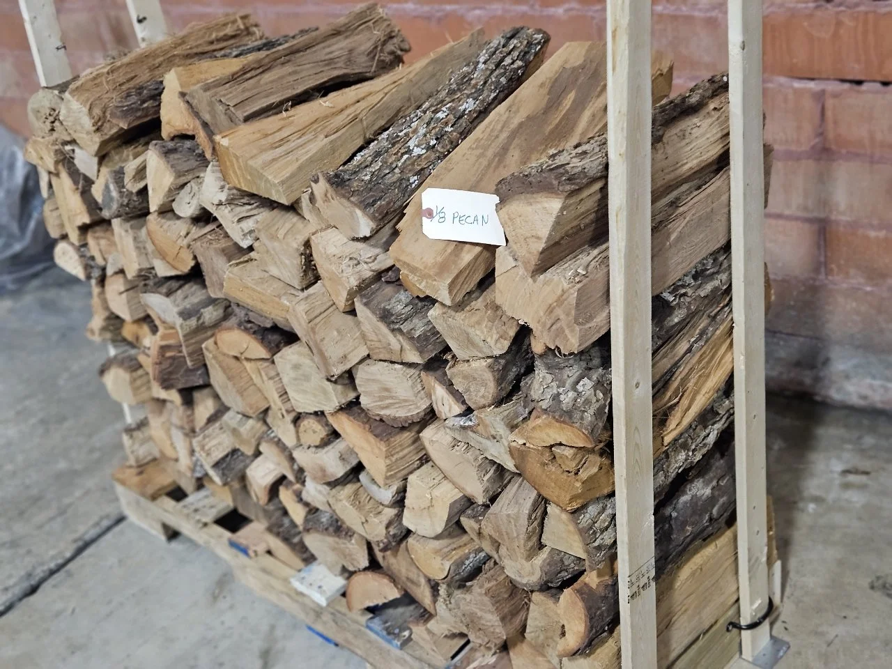 Stacked firewood on a wooden cart with a small handwritten sign labeled 'Pecan' in front of a brick wall.