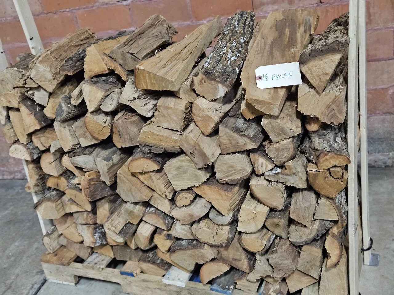 Stack of firewood logs arranged on a wooden cart against a brick wall, with a small white tag that reads '6 Pecan.'