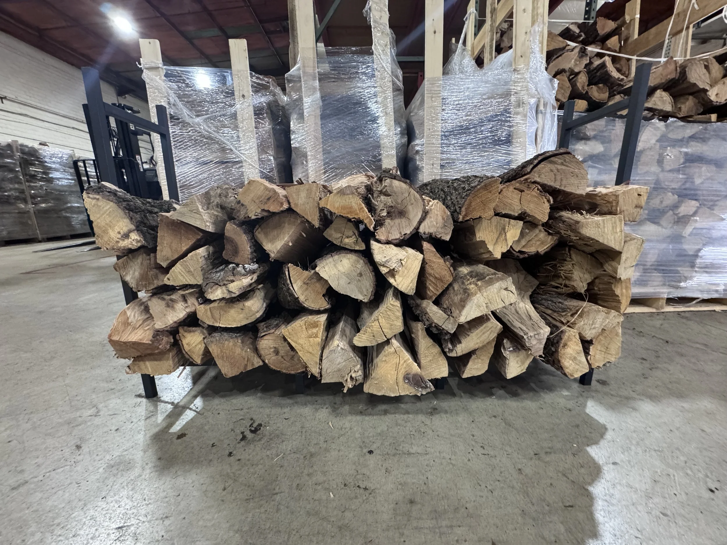 Stacked firewood logs on a metal rack in a warehouse with concrete floor, with shelves and wrapped pallets in the background.