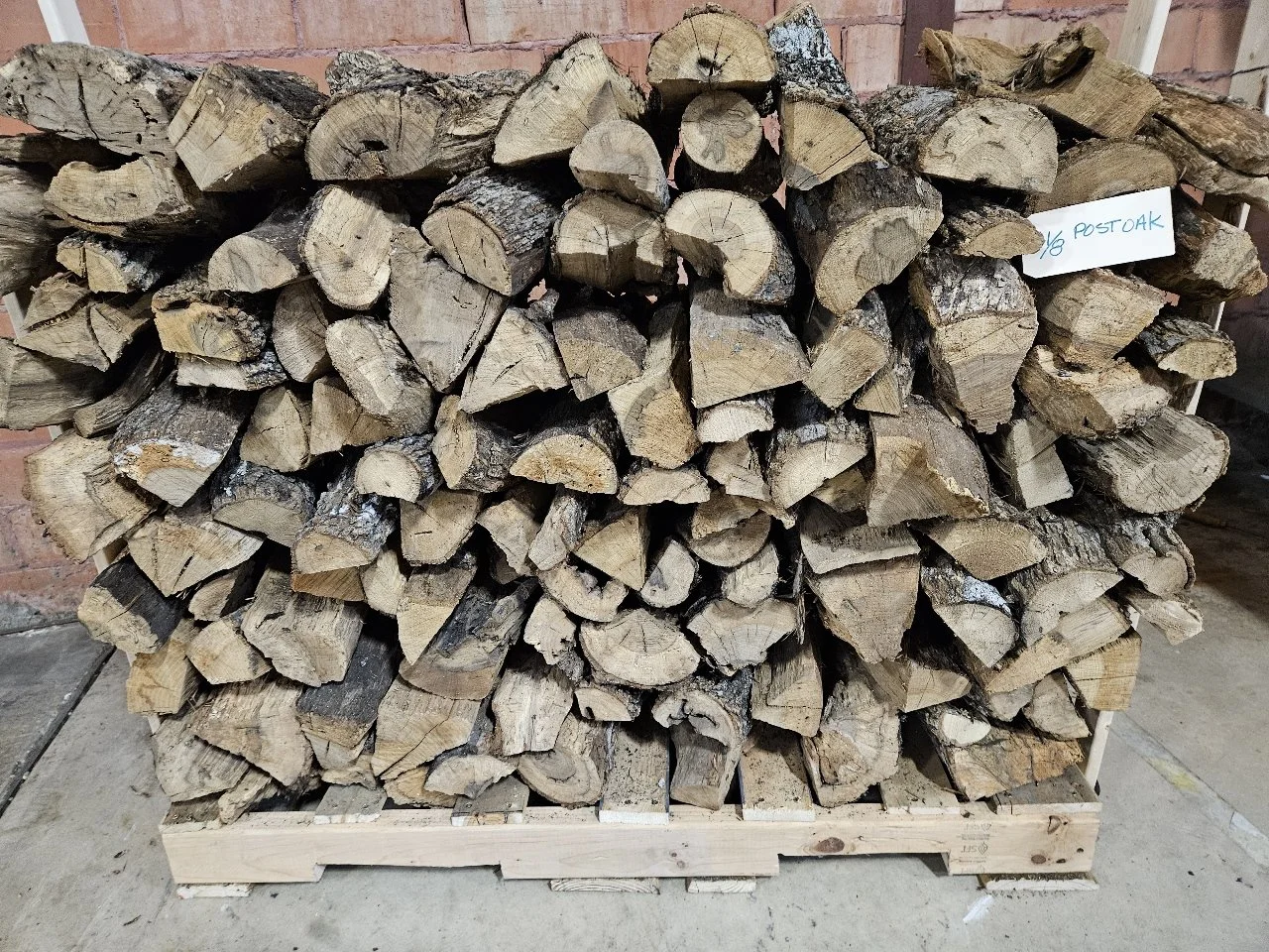Stacked firewood logs on a wooden pallet against a brick wall with a handwritten note attached to the right side.
