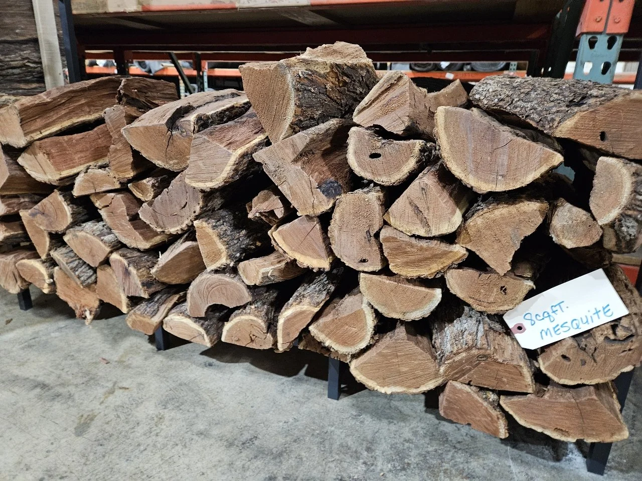 Stack of firewood with a handwritten note labeled "Refr. Mesquite" in a warehouse or storage area.