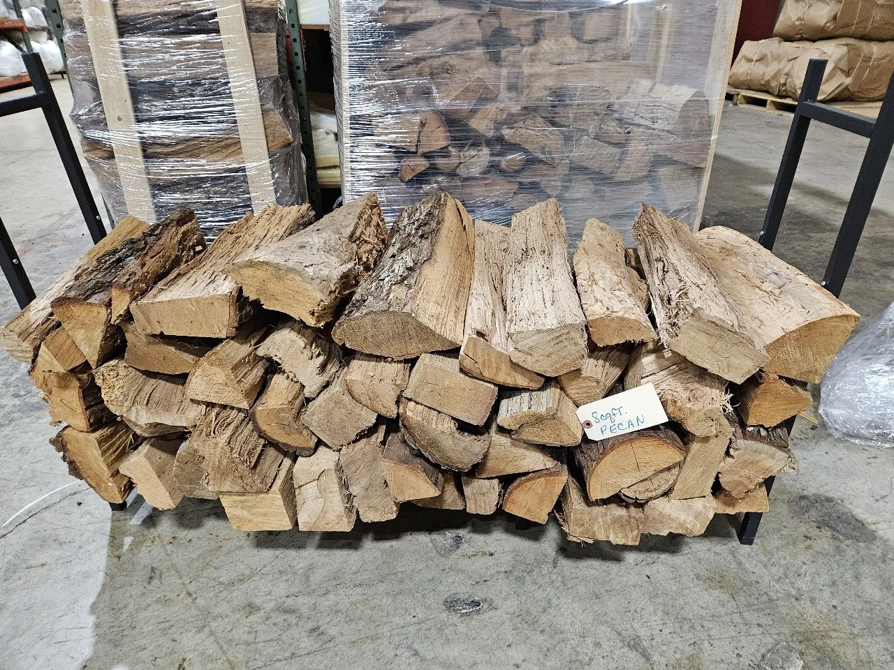 A bundle of firewood logs stacked on a black metal cart, labeled 'Soft Pecan', in a warehouse setting.