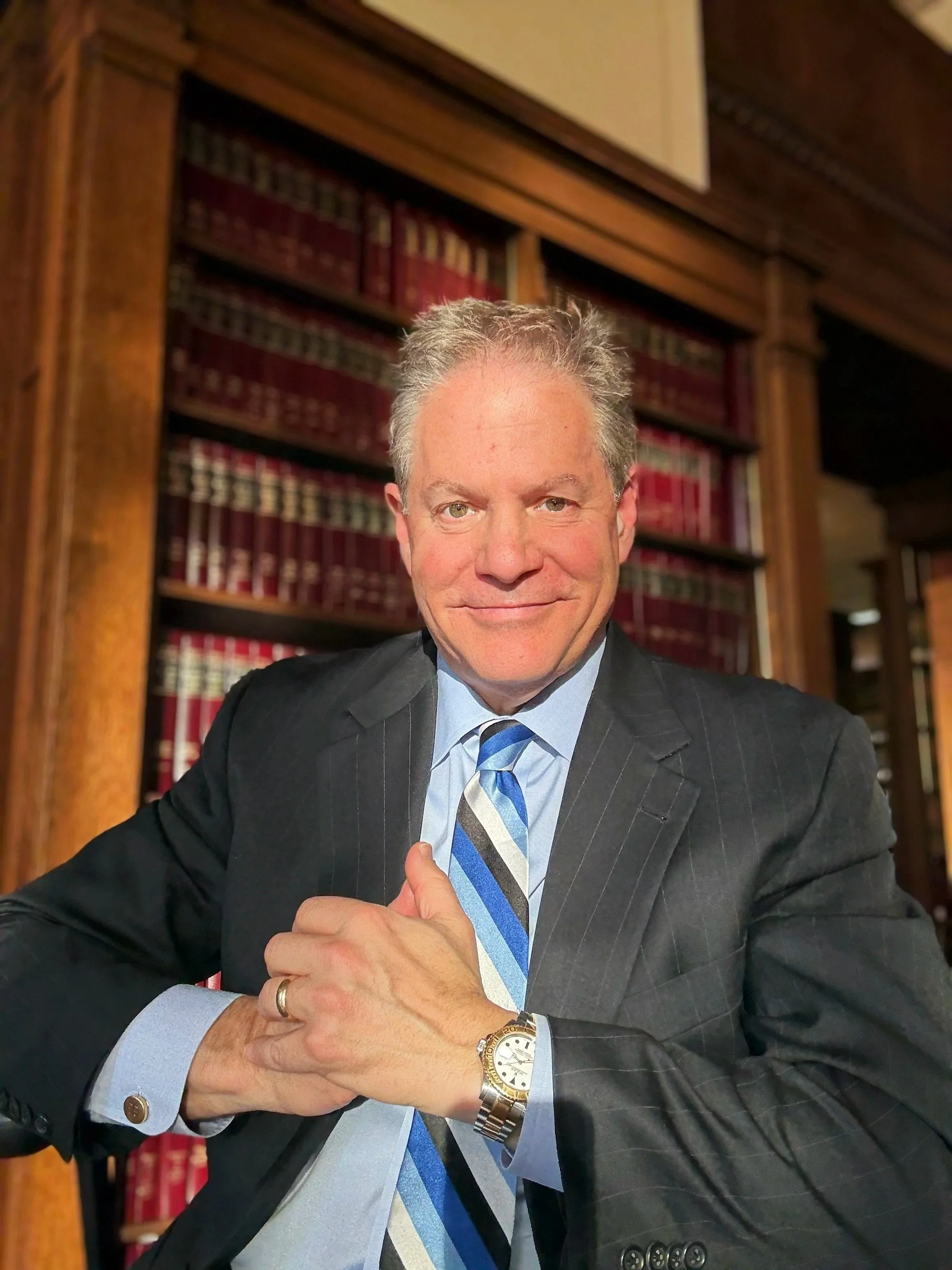A man in a business suit with a striped tie, sitting in a library with wooden bookshelves filled with legal books in the background.