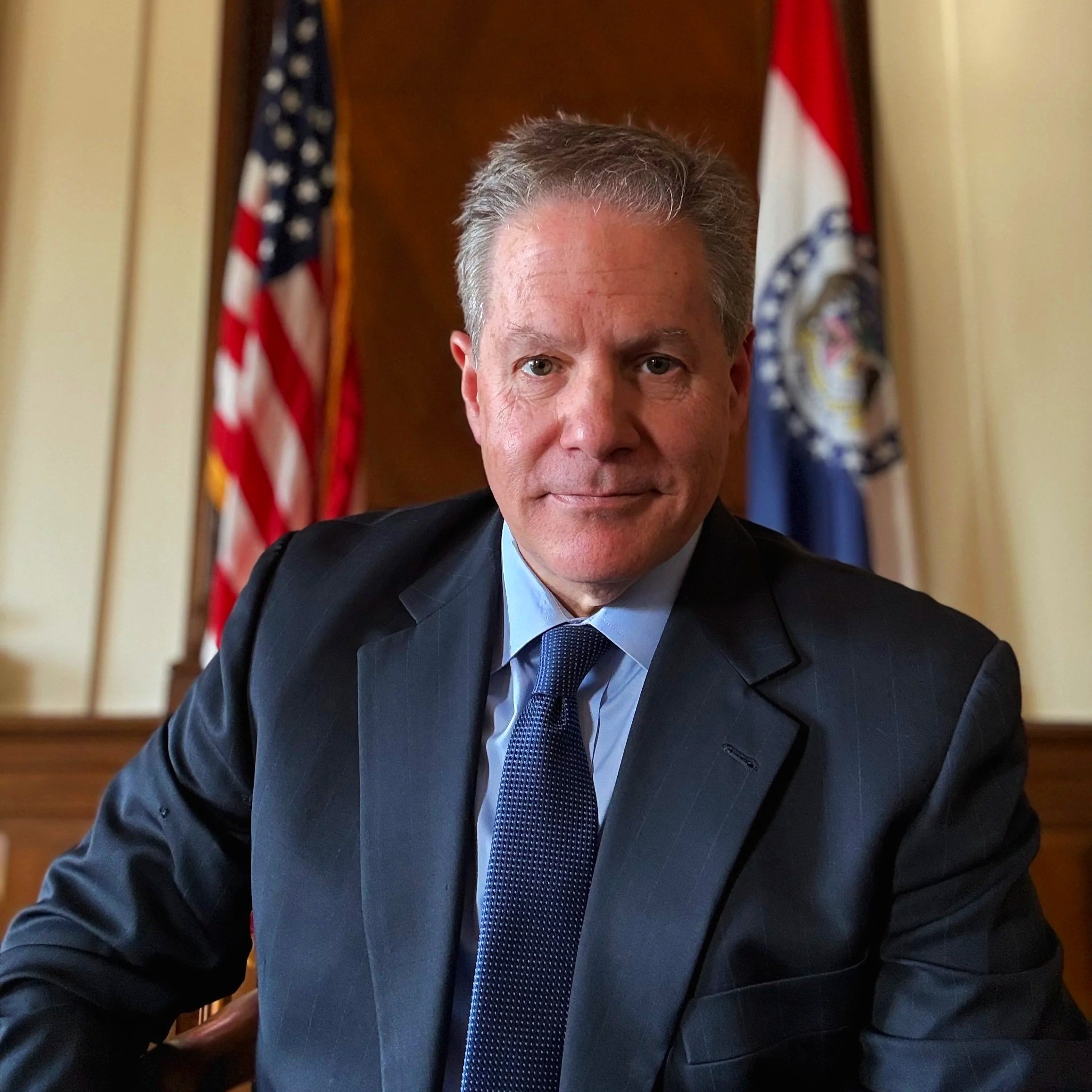 A man in a dark suit and blue tie sitting at a desk with American and state flags in the background.