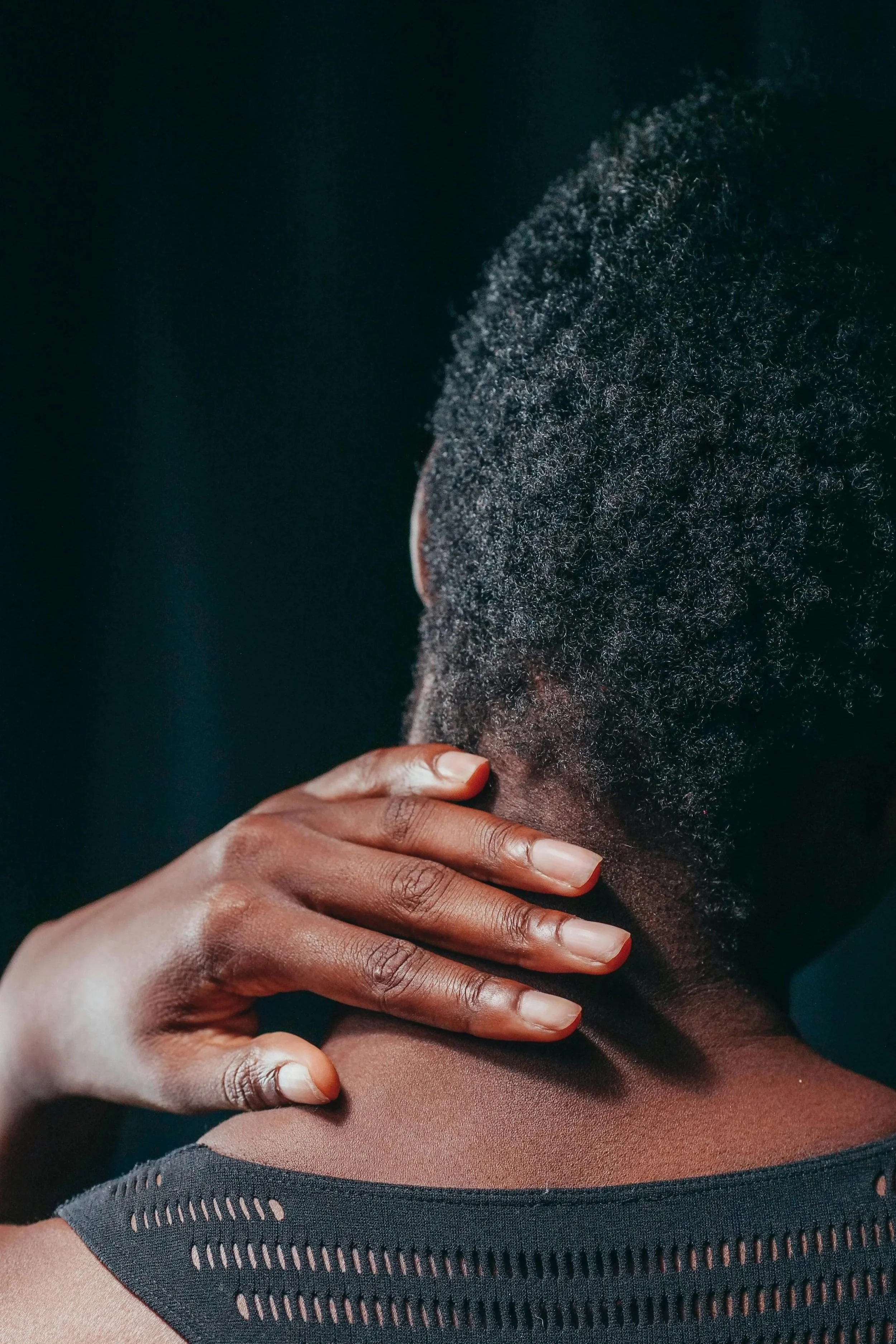 Back of a person with natural textured hair touching their neck, wearing a dark top with cut-out details.