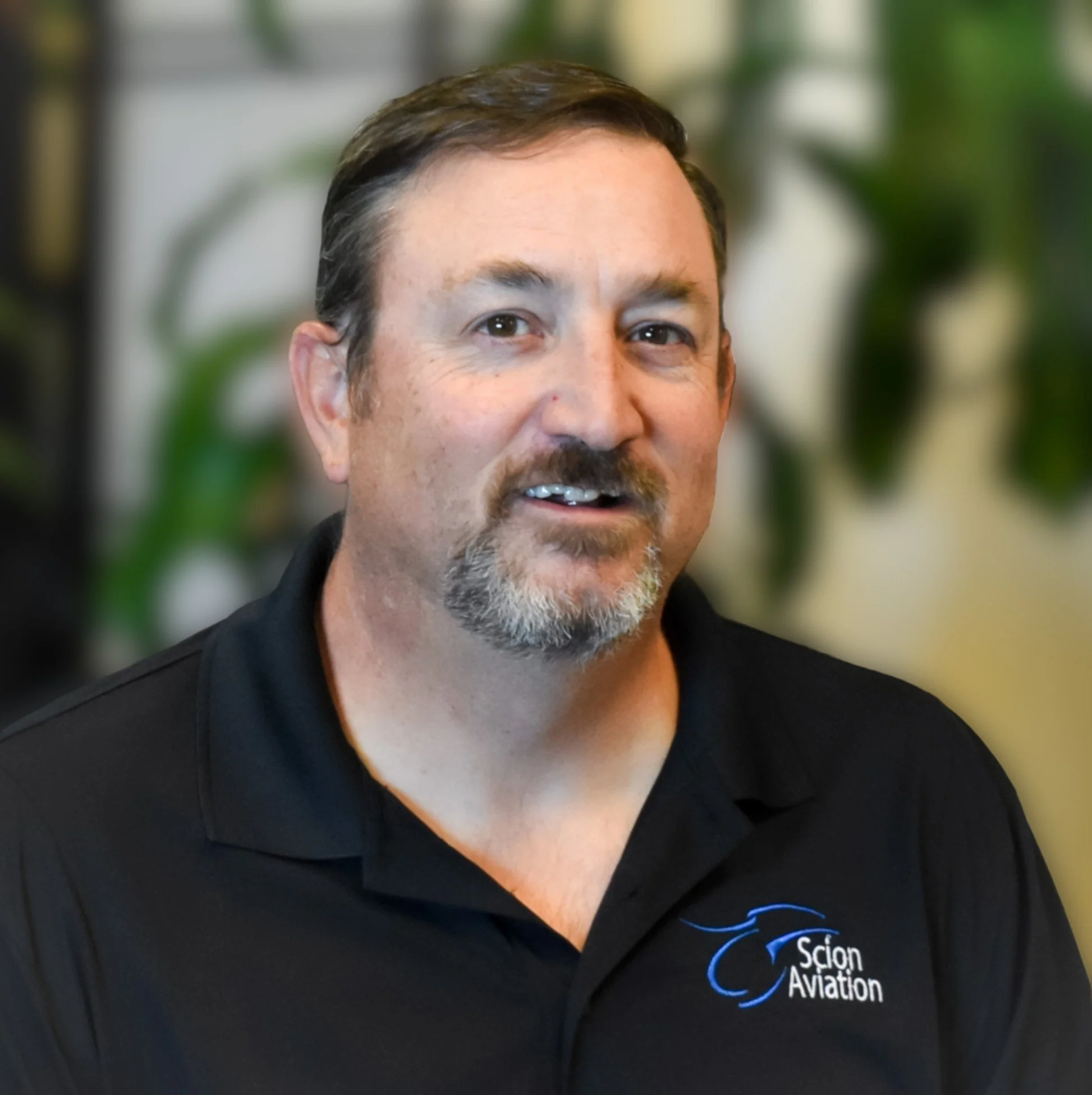 A man with short brown hair, a goatee with gray, wearing a black polo shirt with a blue logo that says 'Scion Aviation', standing in front of blurred green plants.