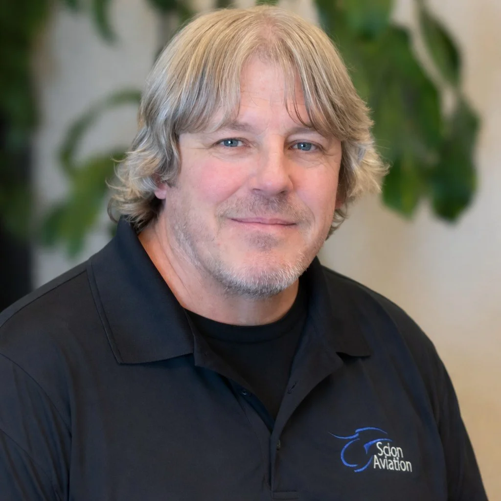 A middle-aged man with shoulder-length gray hair and beard, wearing a black polo shirt with a logo that says 'Salon Aviation,' smiling in front of a blurred background of green plants.