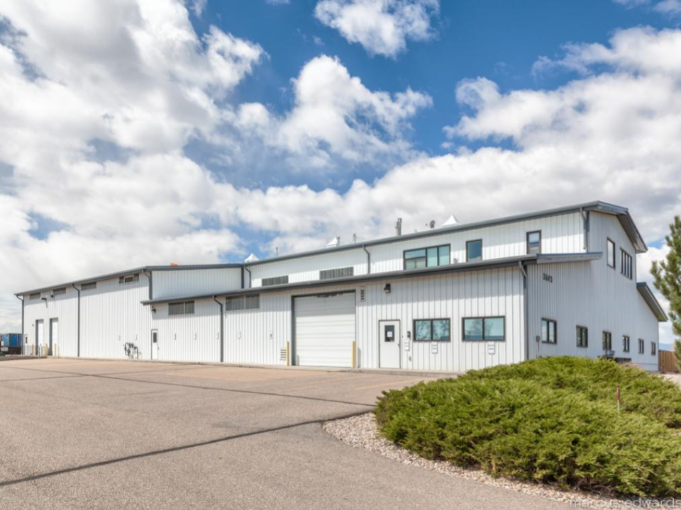 Large white industrial or commercial building with multiple windows and a garage door, set in a sunny area with a partly cloudy sky and greenery in the foreground.