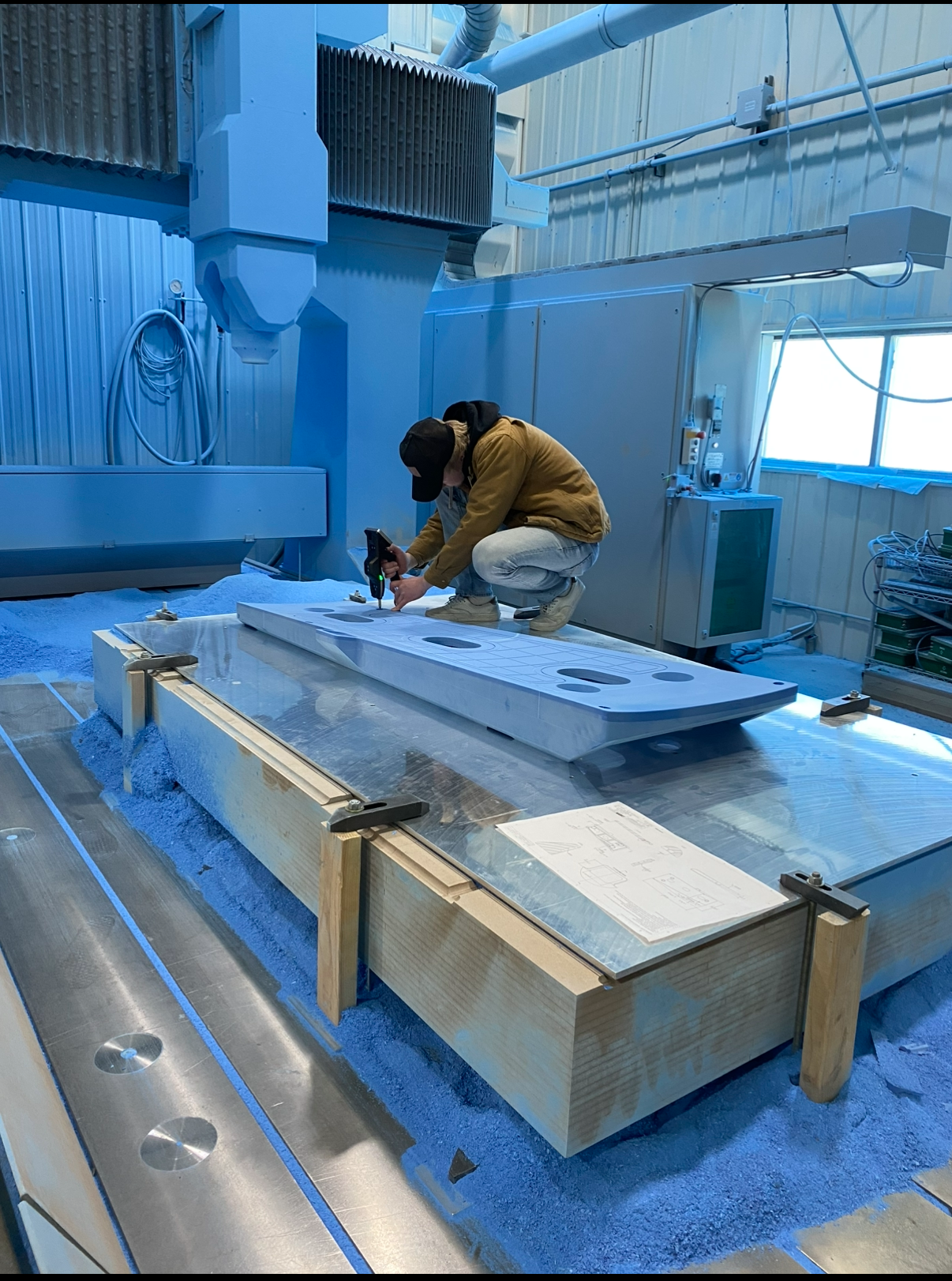 A person wearing a brown jacket, black cap, and tan boots kneeling on a large worktable, using a handheld device to measure or examine a long, flat, white object with circular cutouts inside a manufacturing or industrial setting with blue lighting.